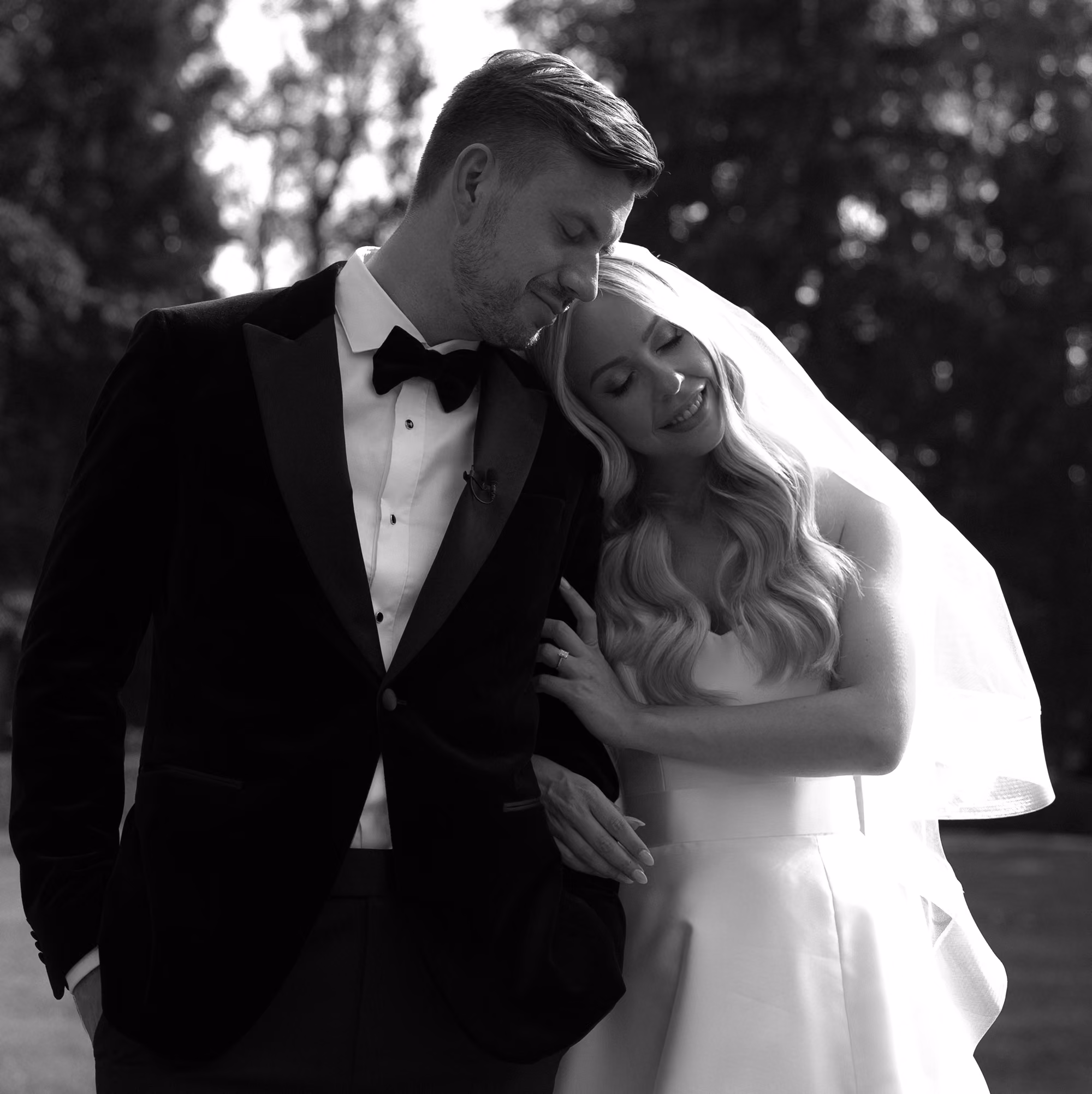 Bride in veil leaning her head on groom’s shoulder as he smiles in tuxedo outdoors.