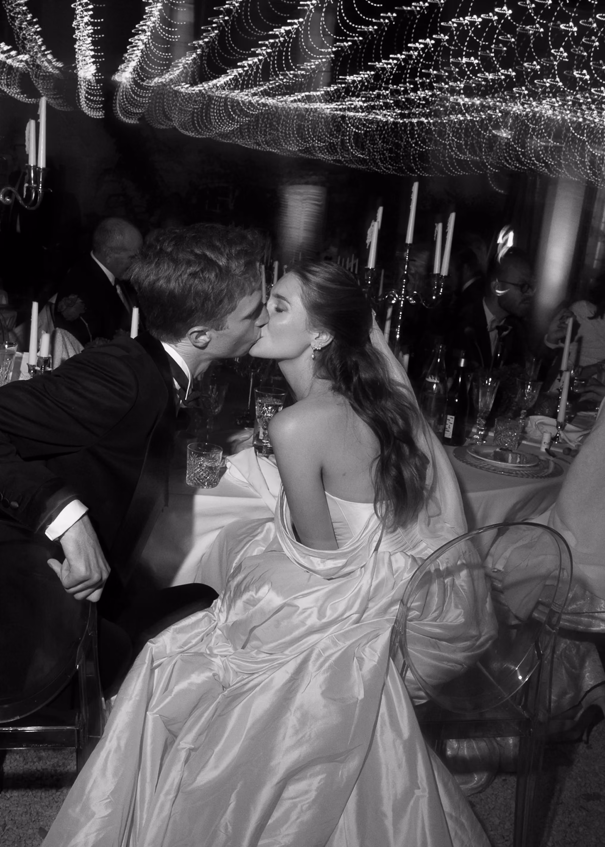 Bride and groom kissing at their wedding reception with string lights overhead in black and white.