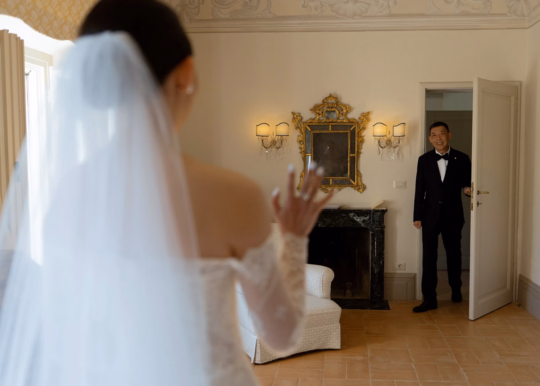 Bride in wedding dress waving to smiling groom entering room through a door.