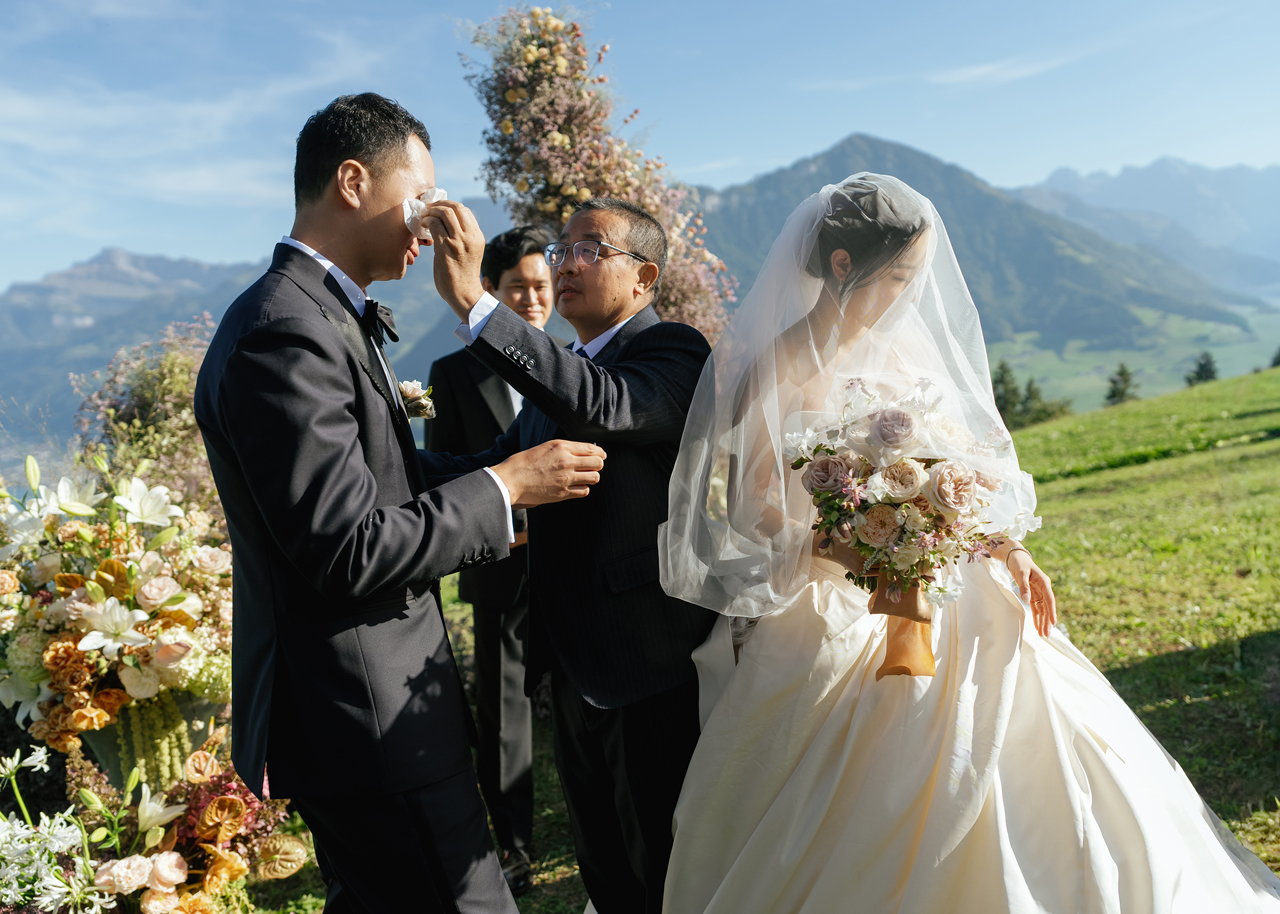 Bride in wedding gown and veil holding bouquet walks arm-in-arm with older man in tuxedo outdoors.