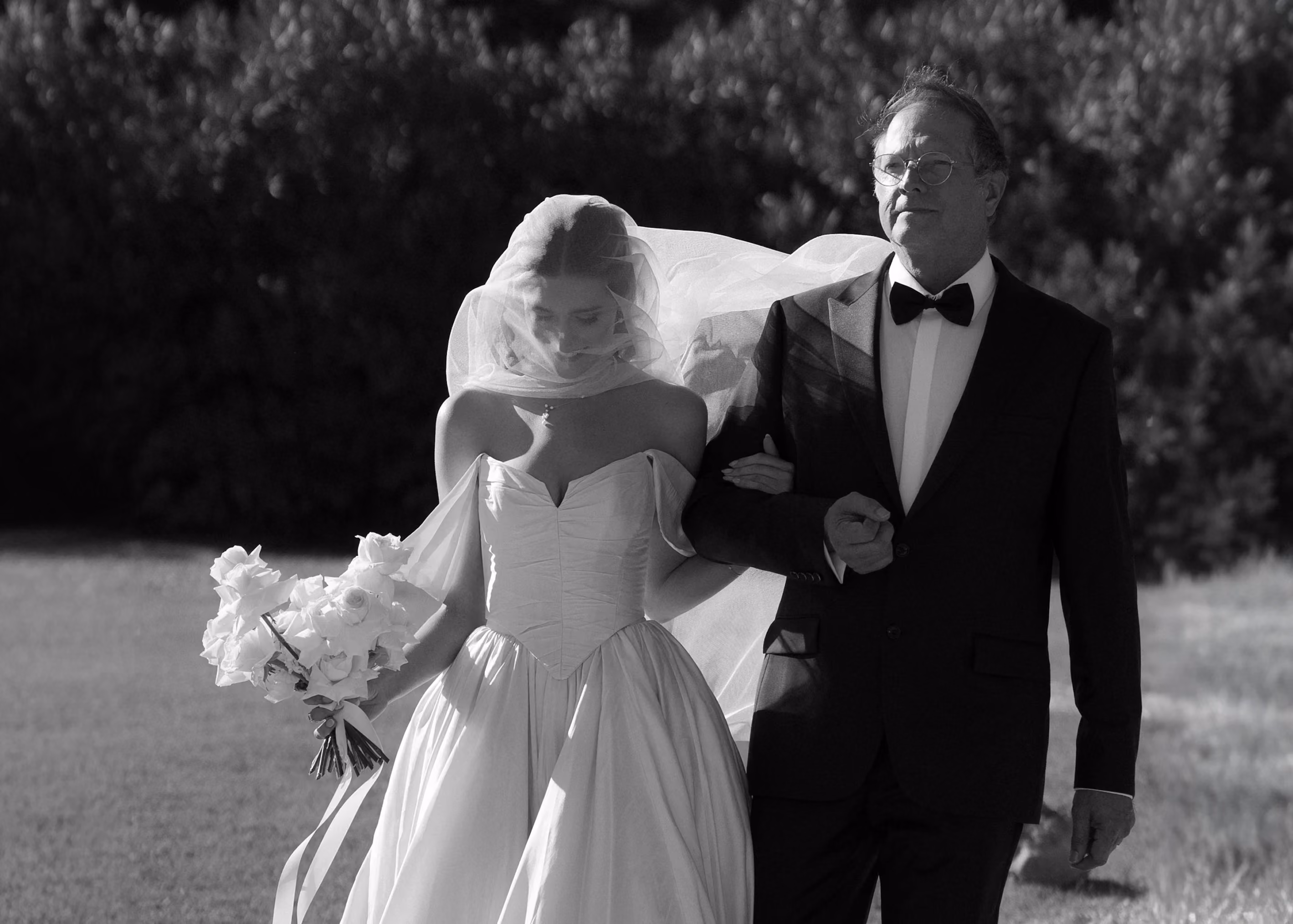 Bride in wedding gown and veil holding bouquet walks arm-in-arm with older man in tuxedo outdoors.