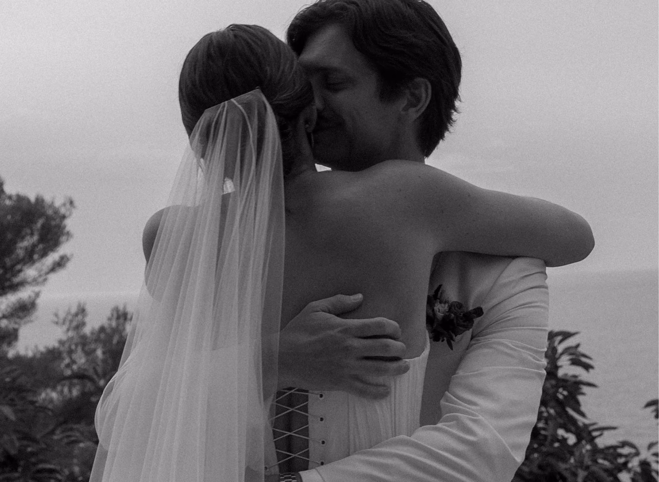 Black-and-white photo of a bride and groom embracing outdoors near trees and water.
