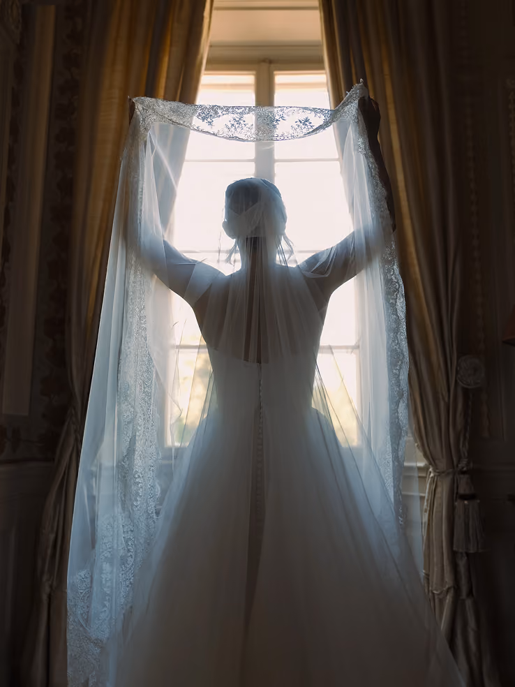 Bride in wedding dress holding lace veil up in front of a bright window with curtains.