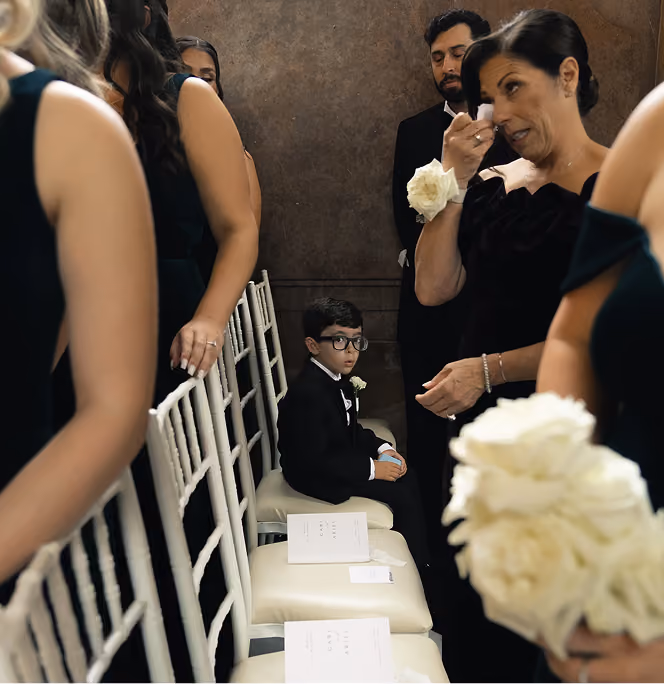 A young boy in a tuxedo sits among adults in formal attire, while a woman wipes away a tear.
