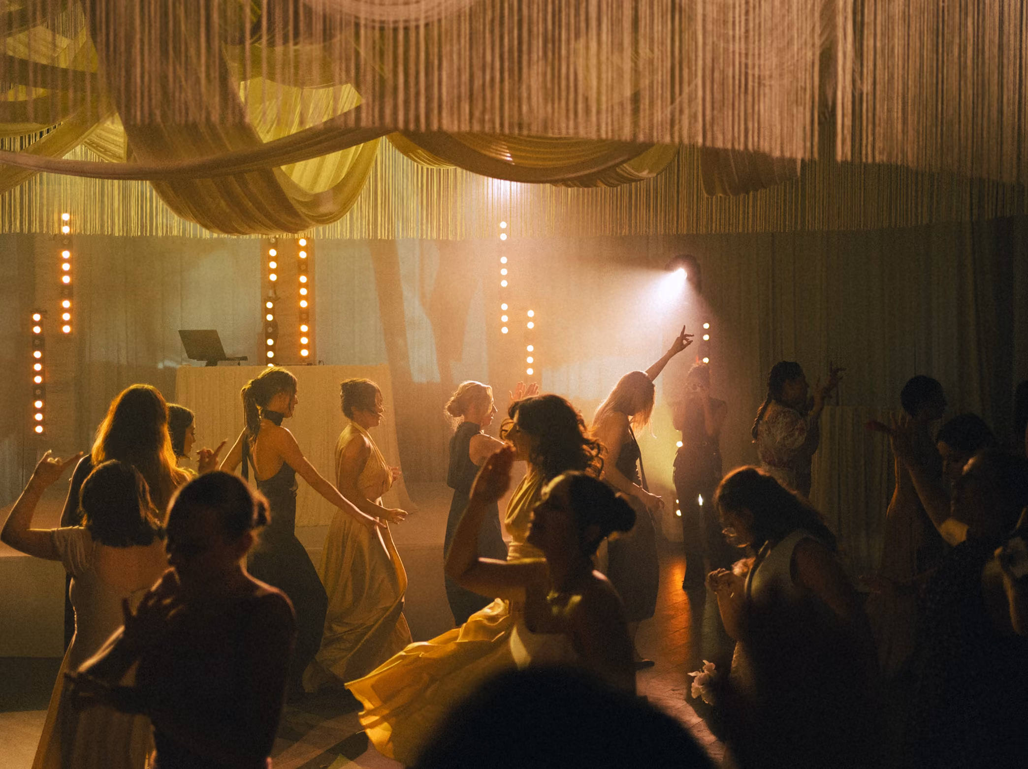 Women in elegant dresses dancing under warm lighting and draped decorations in a festive indoor setting.