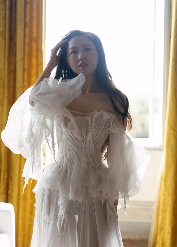 Woman in a white, sheer, ruffled dress standing indoors with yellow curtains and a bright window behind her.