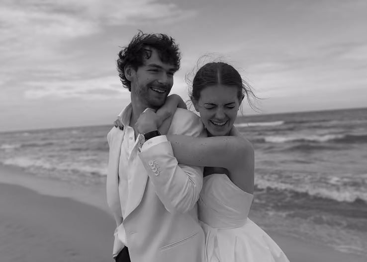 Smiling couple in wedding attire embracing and laughing on a beach with ocean waves behind them.