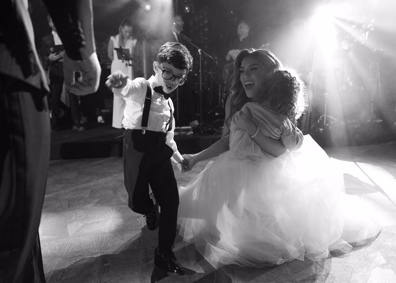 Boy wearing glasses and suspenders dancing with woman in a gown holding a small child in a bright event setting.