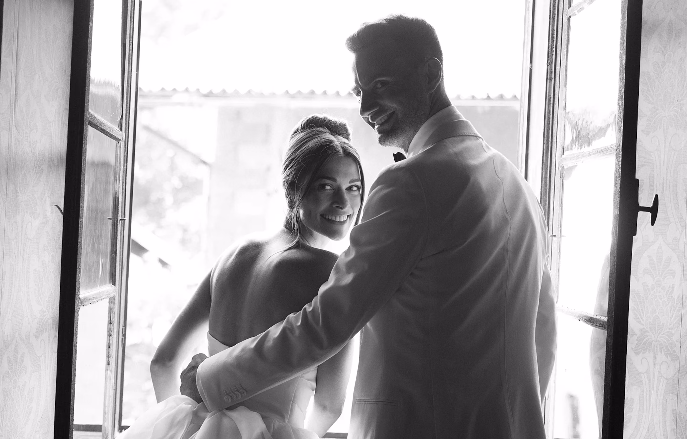 Smiling bride and groom embracing near an open window with bright light outside.
