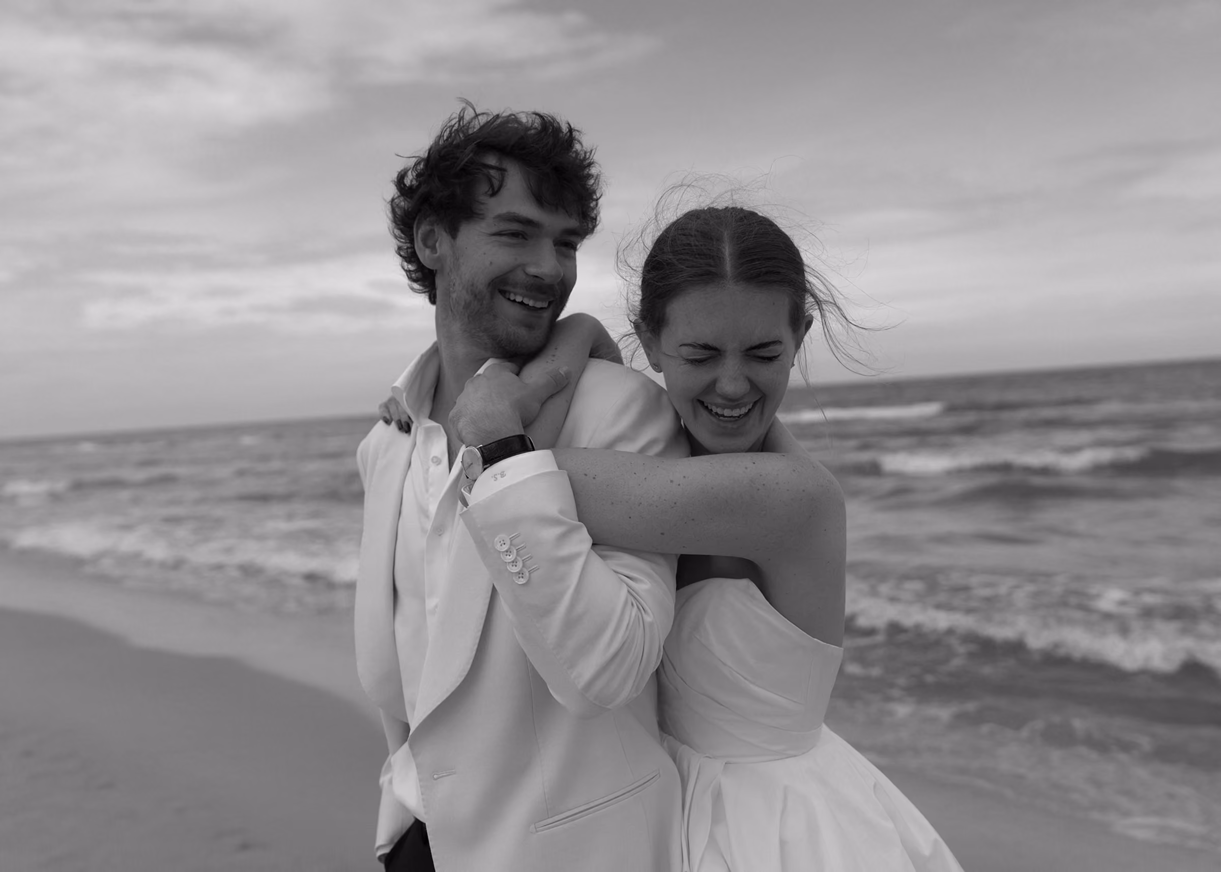 Couple in wedding attire laughing and embracing on a beach with waves in the background.