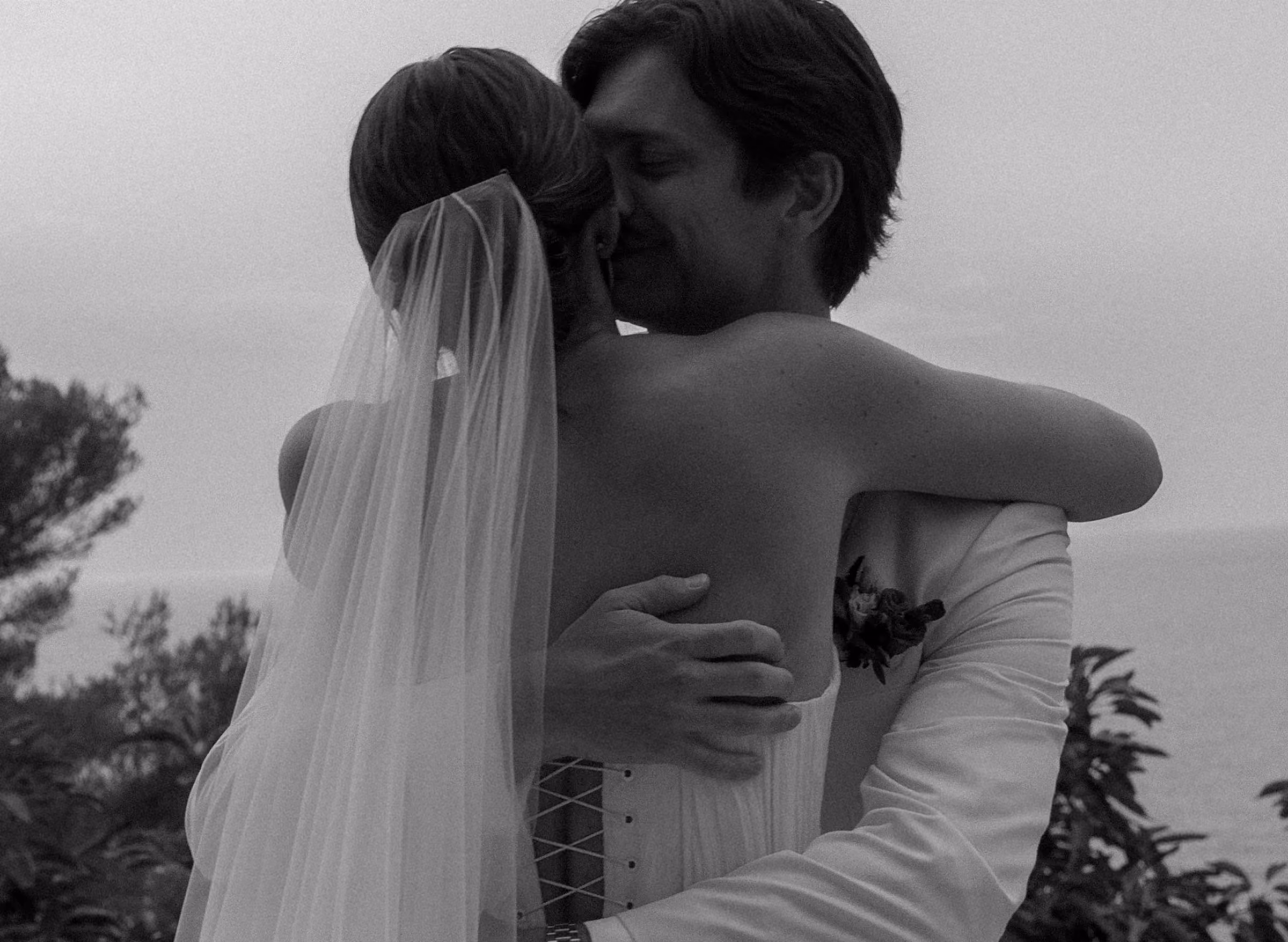 Black and white photo of a bride and groom embracing outdoors near trees and water.
