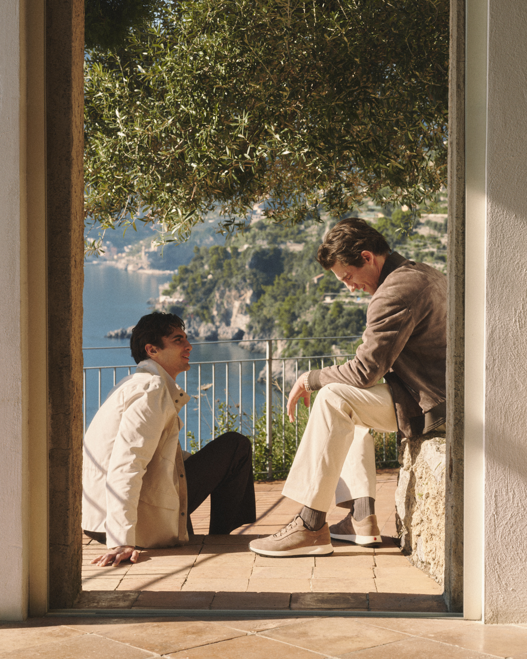 Two men sit on a terrace framed by a doorway overlooking a coastal landscape with trees and cliffs.