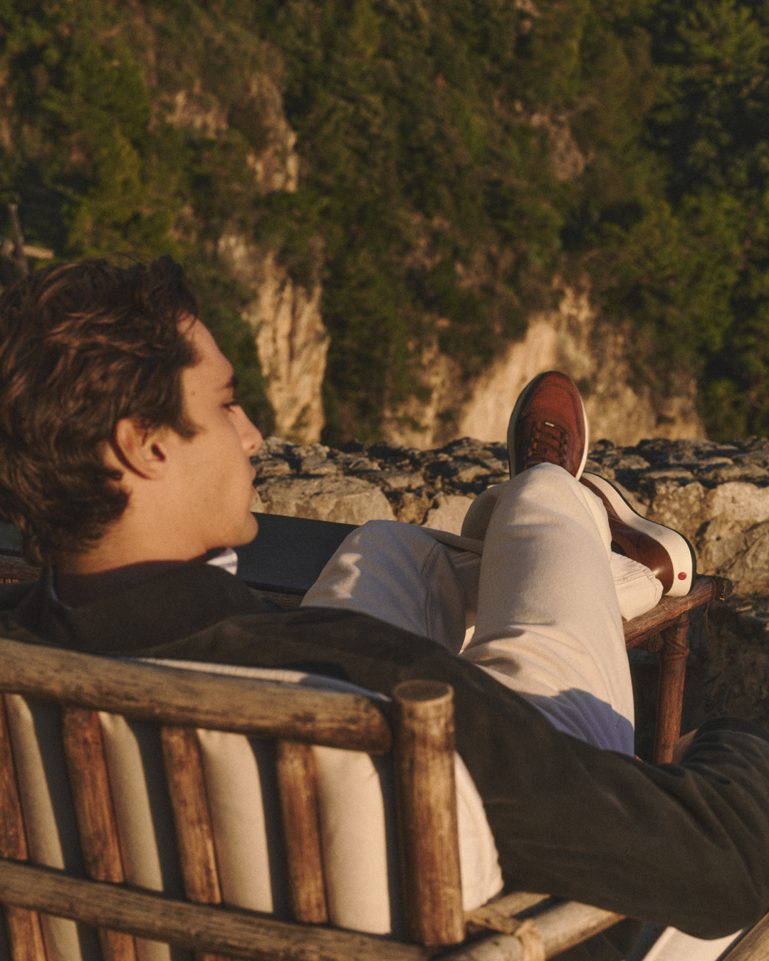 Man reclining on a wooden chair outdoors overlooking rocky cliffs and green trees.