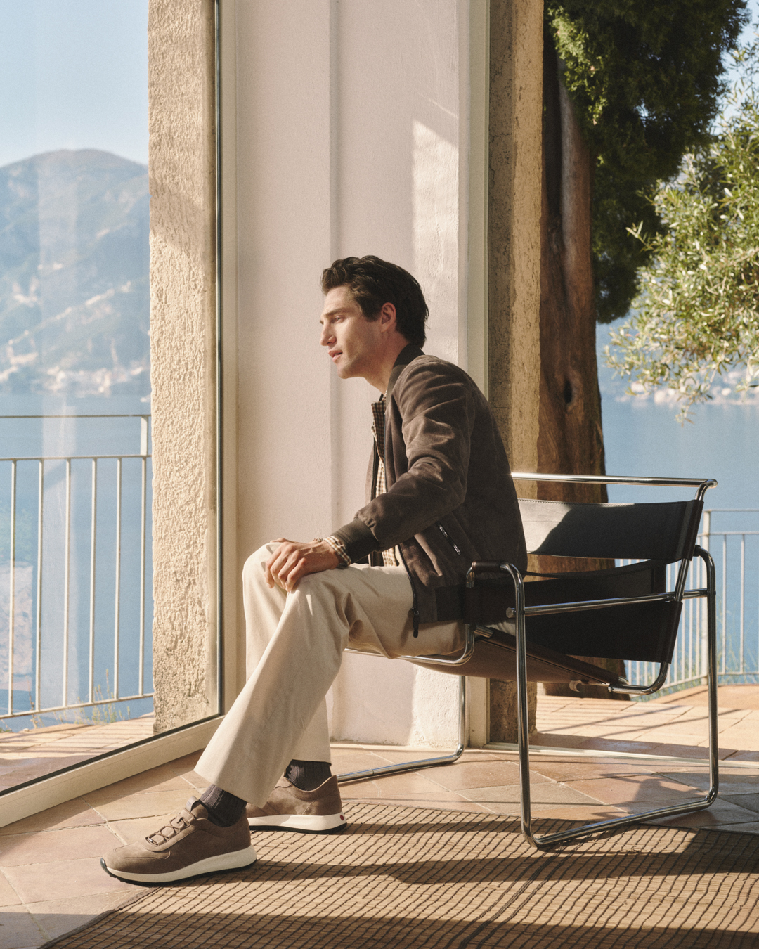 Man in brown jacket and beige pants sitting on modern metal chair by window overlooking mountains.