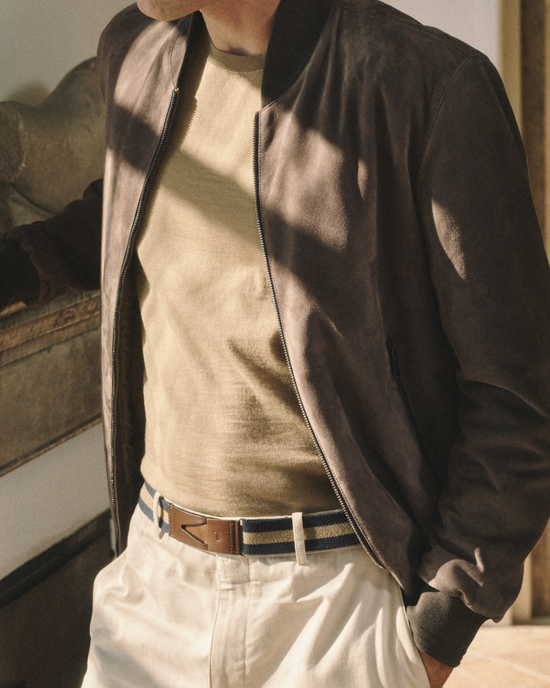 Man wearing a brown suede jacket, beige sweater, white pants, and striped belt.