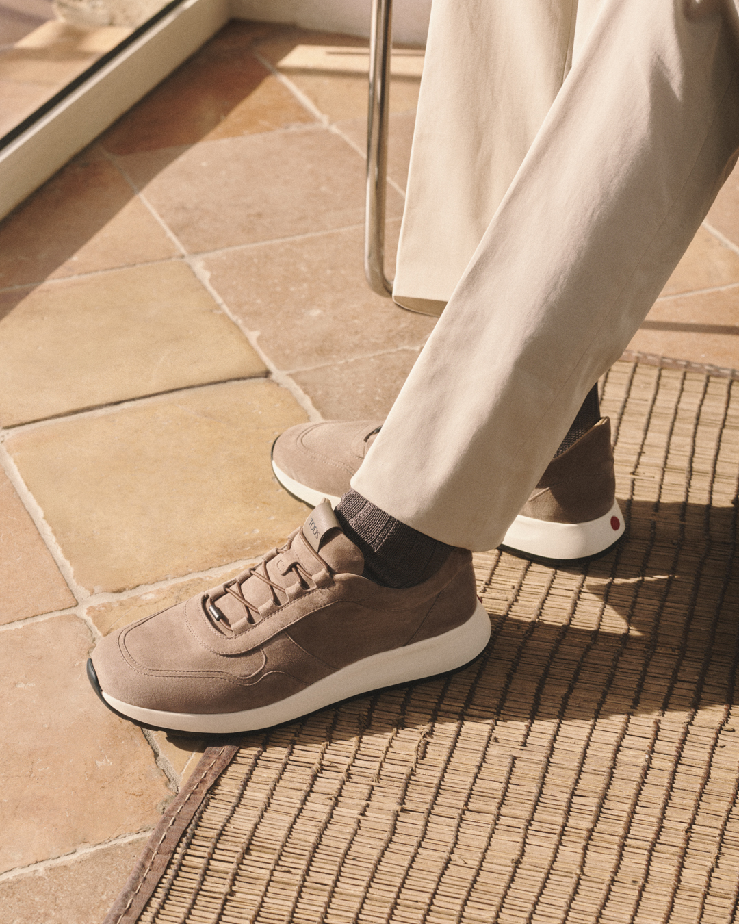 Person wearing taupe suede sneakers with white soles and beige pants sitting on a woven mat indoors.
