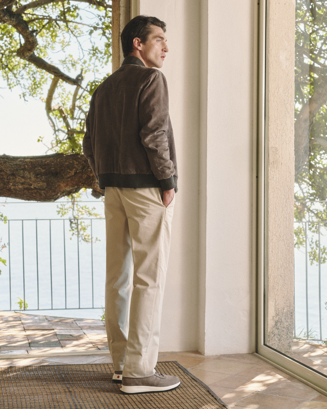 Man in brown jacket and beige pants standing indoors near glass door looking outside with hands in pockets.