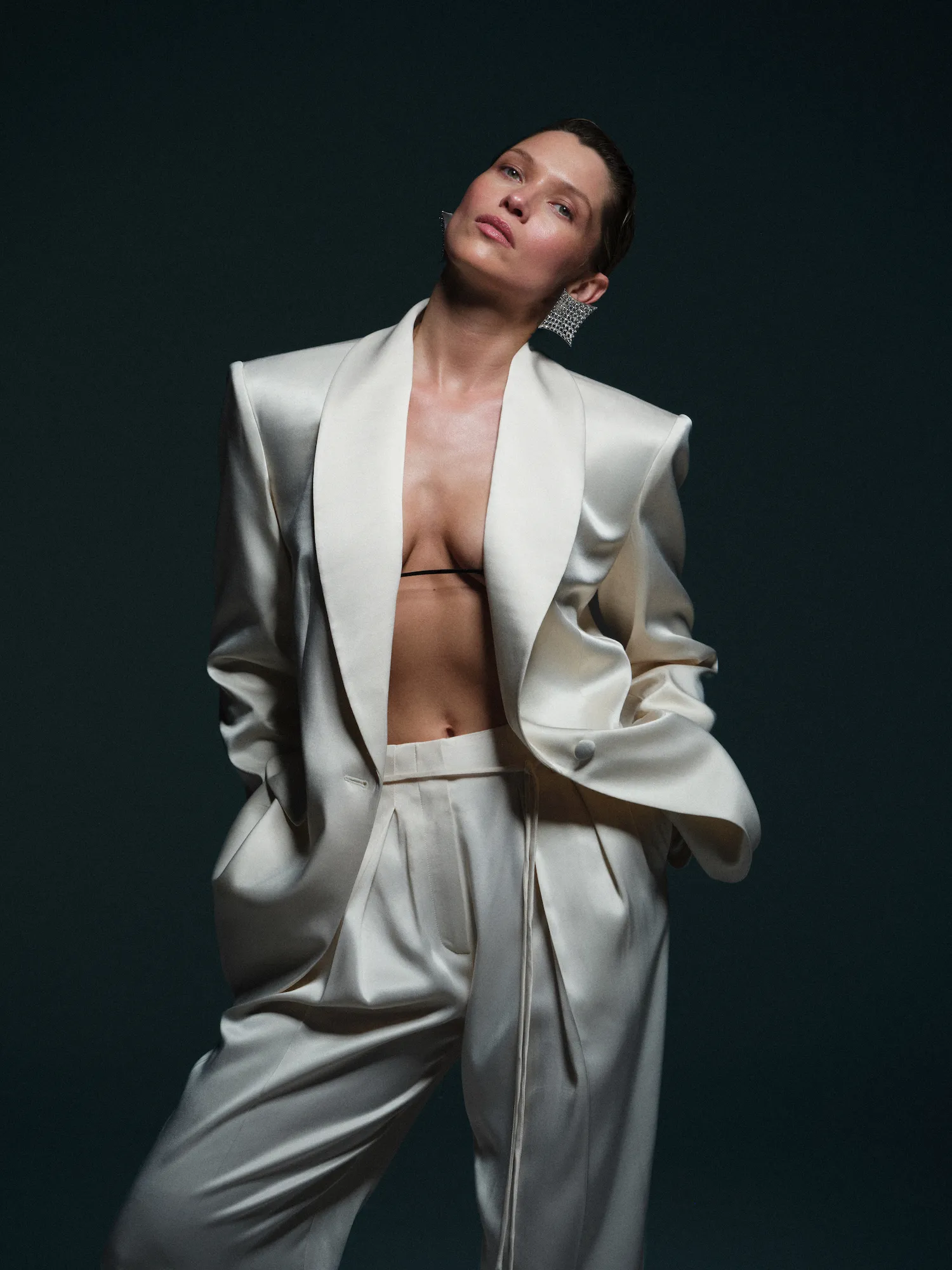 Woman in loose satin cream suit with no shirt underneath, large earrings, and hands in pockets.