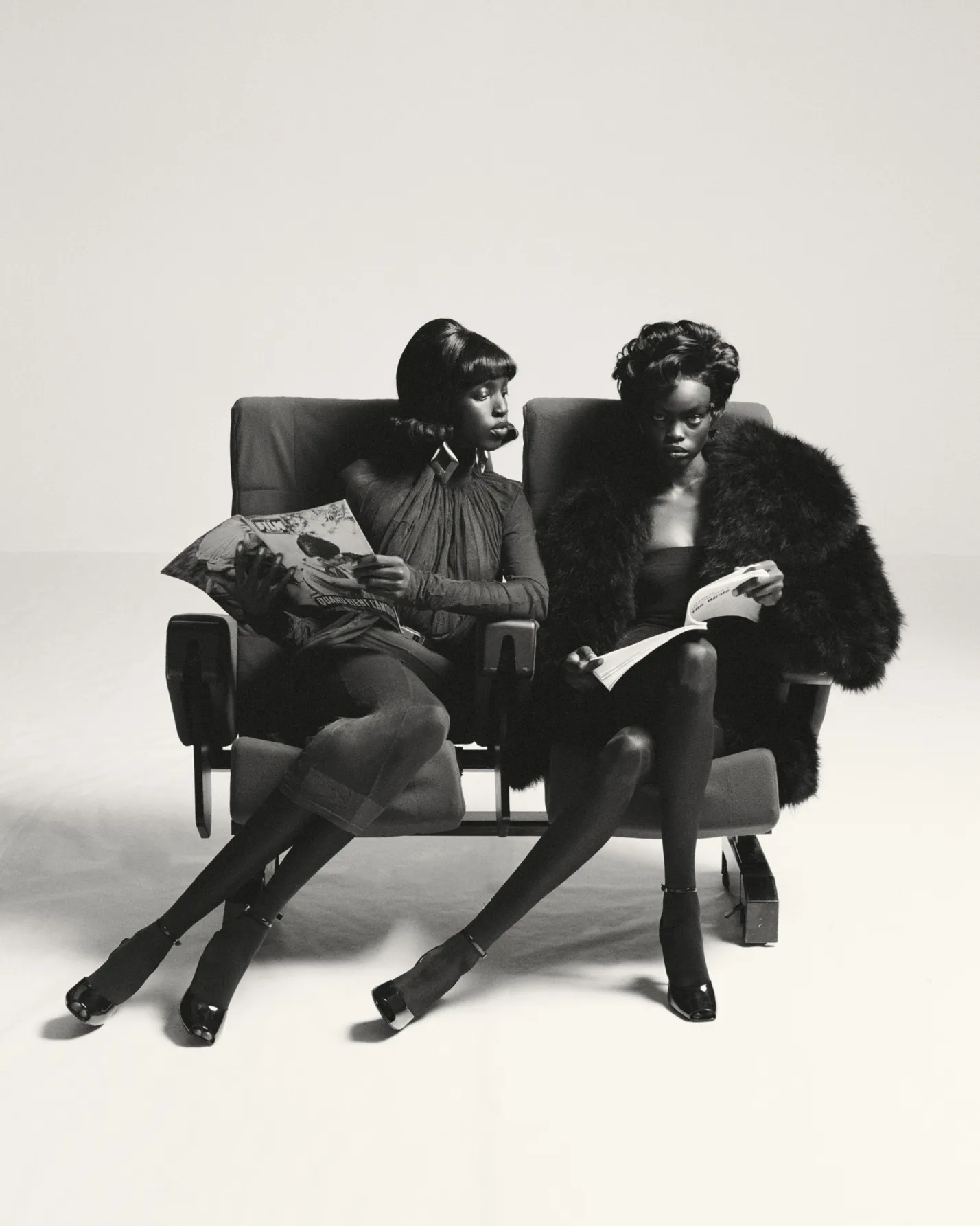 Two women seated side by side in theater chairs, reading magazines in black and white photo.