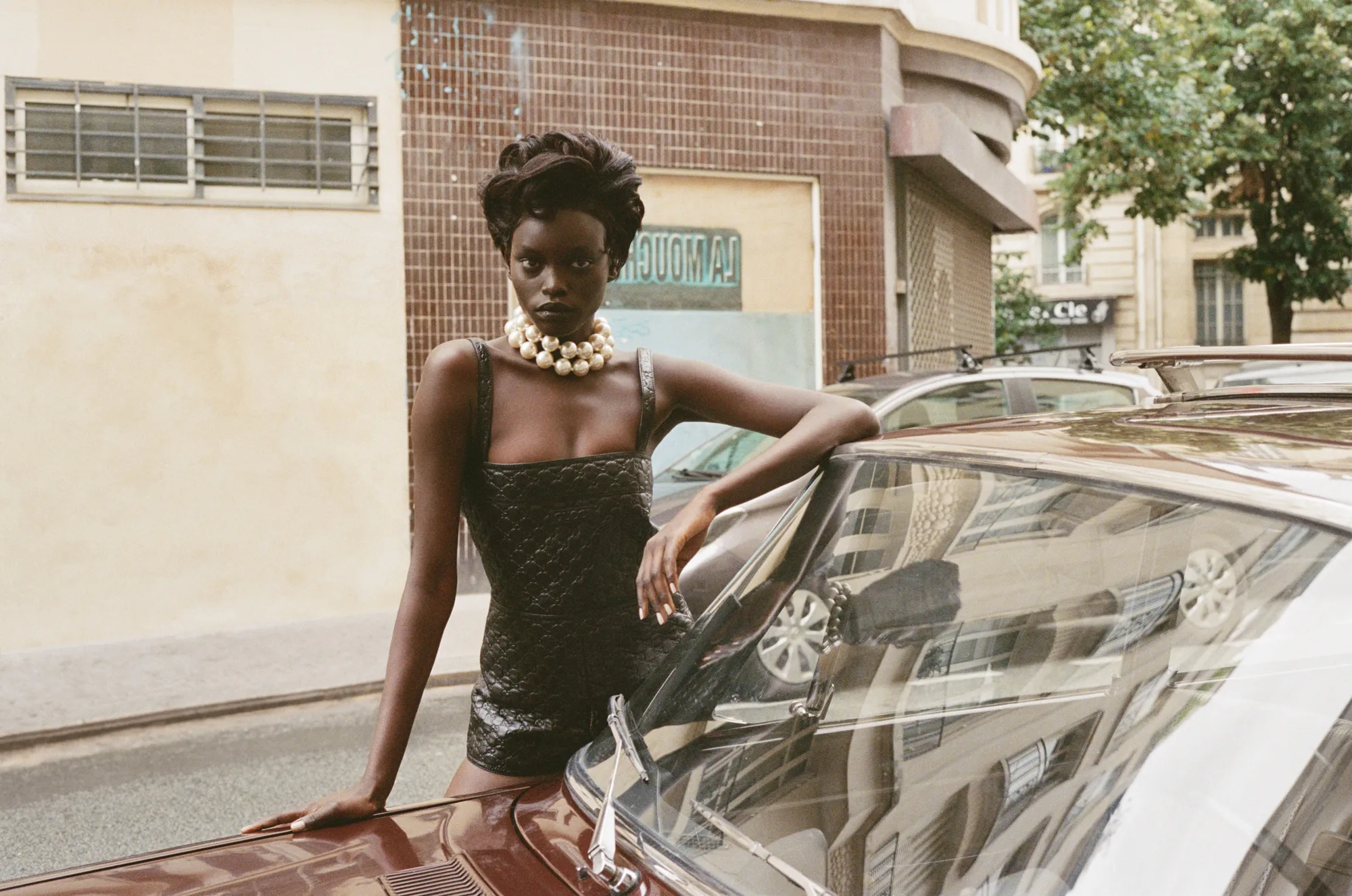 Woman in a black leather outfit and chunky pearl necklace leaning on a vintage brown car on a city street.