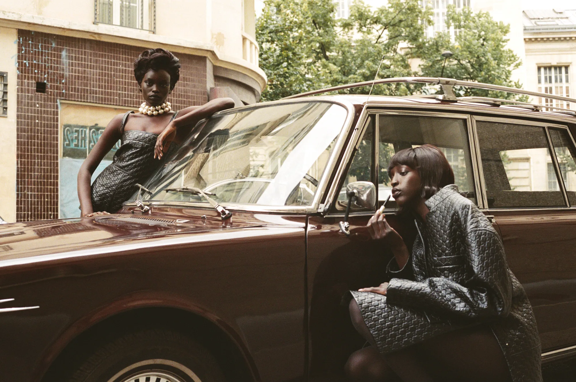 Two women in black textured outfits posing with a vintage brown car on a city street.