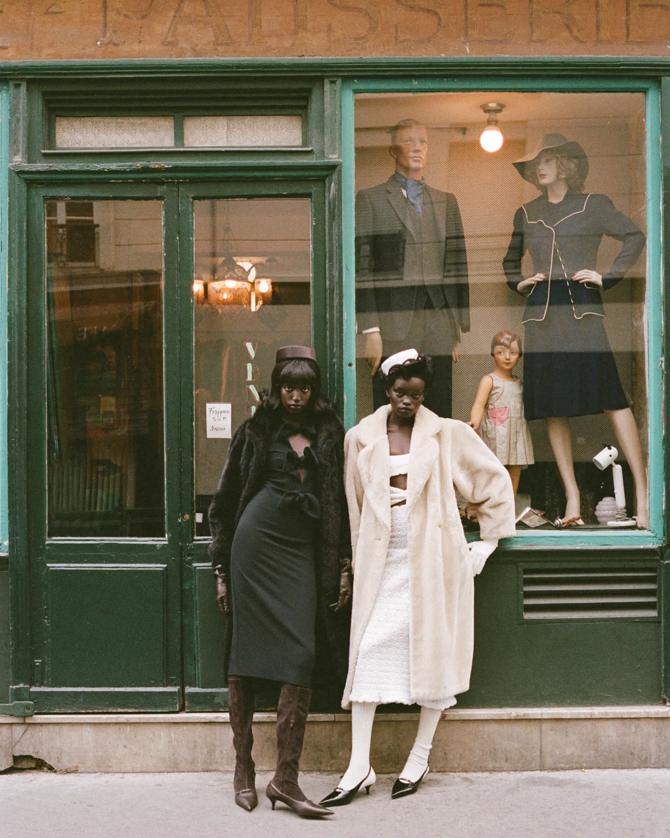 Two women in vintage-style coats and hats posing outside a green store window with mannequins inside.