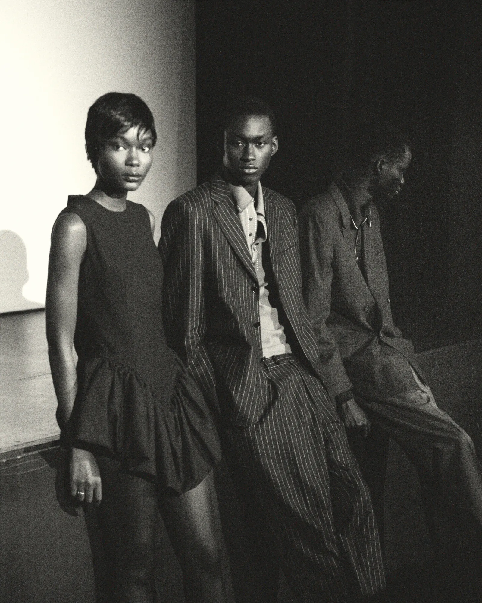 Three stylishly dressed individuals posing in dim light, one woman and two men in dark modern outfits.
