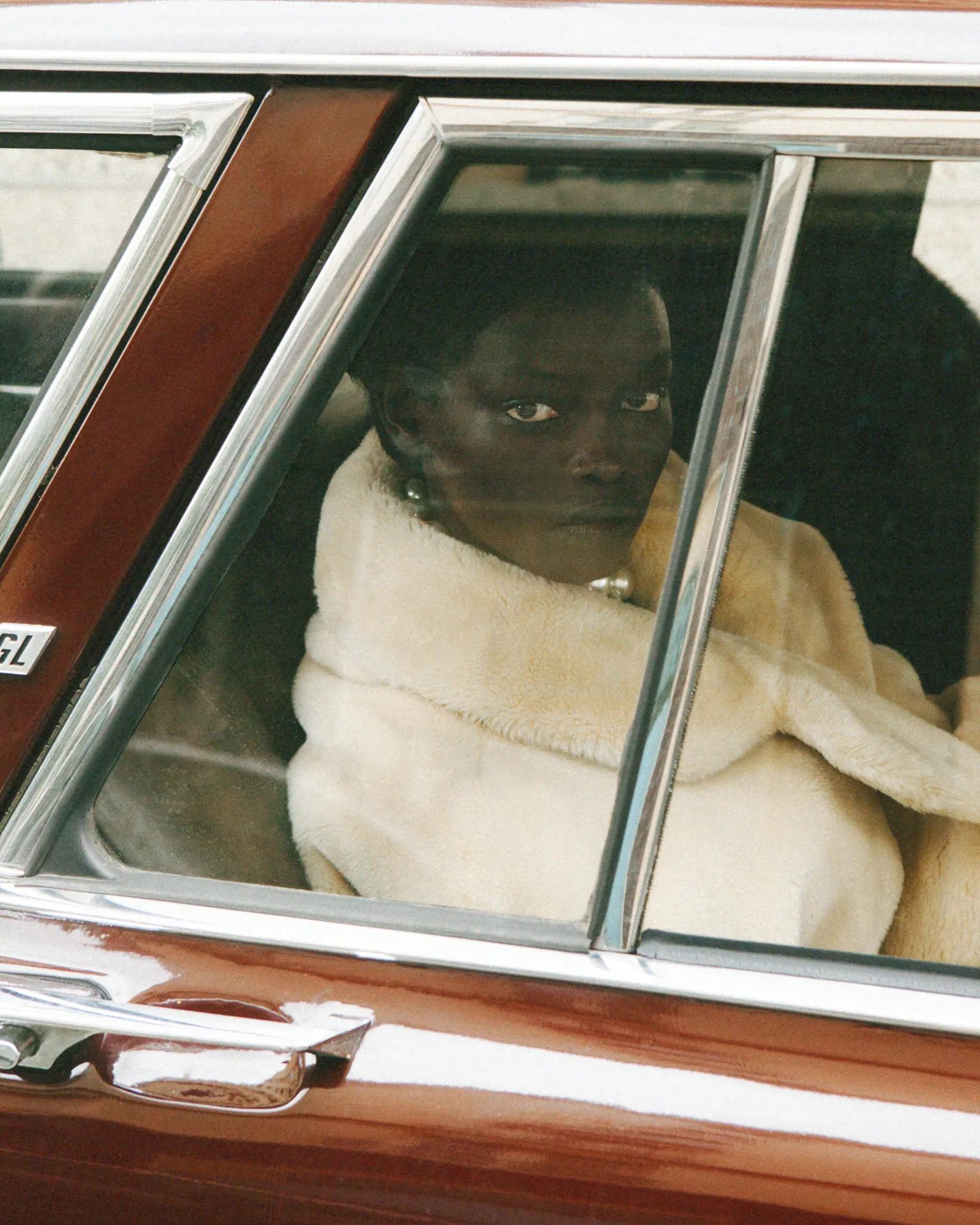 Person in a white fur coat and pearl necklace sitting in a vintage brown car, looking out the window.