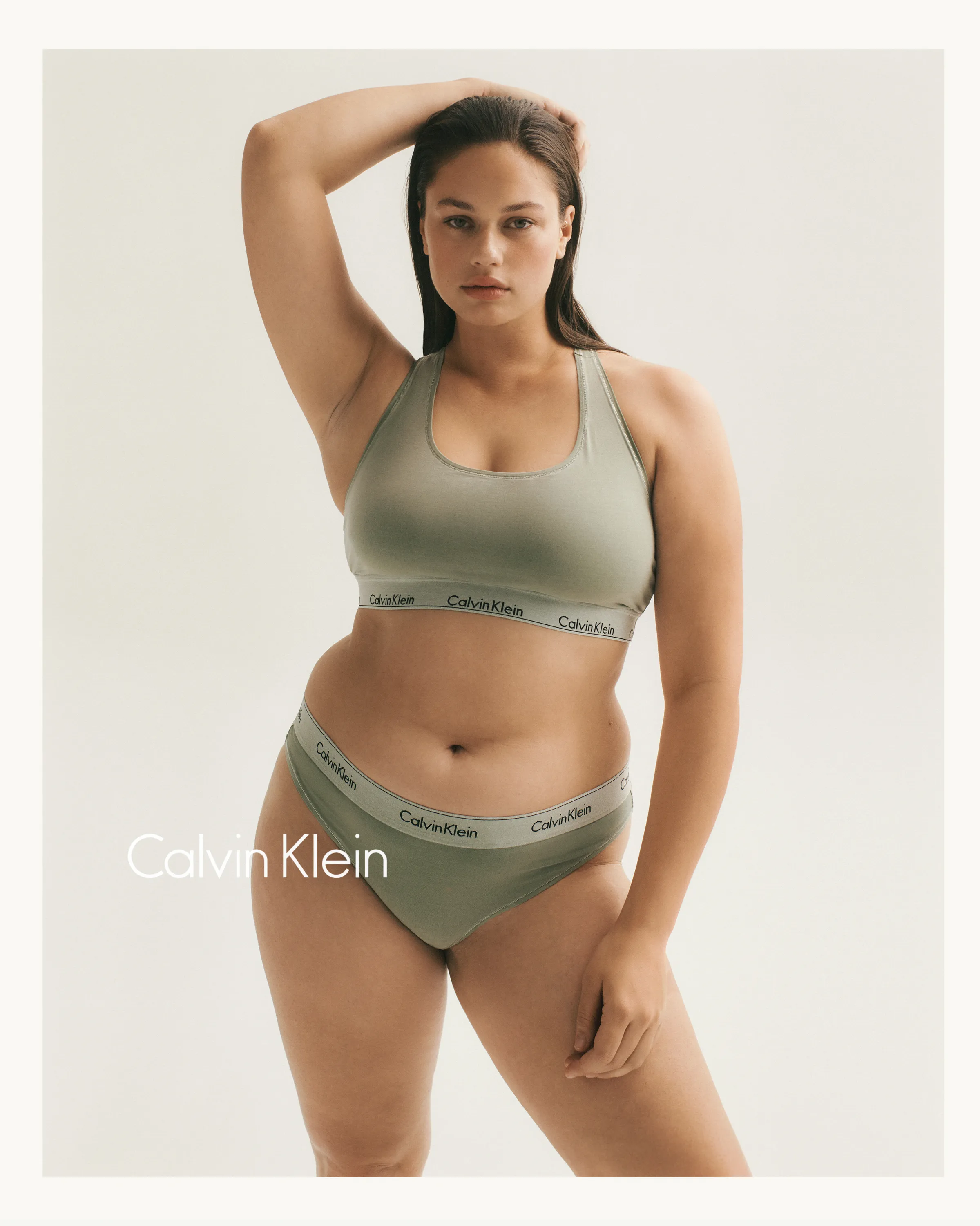 Woman wearing sage green Calvin Klein sports bra and matching underwear posing with arm raised.