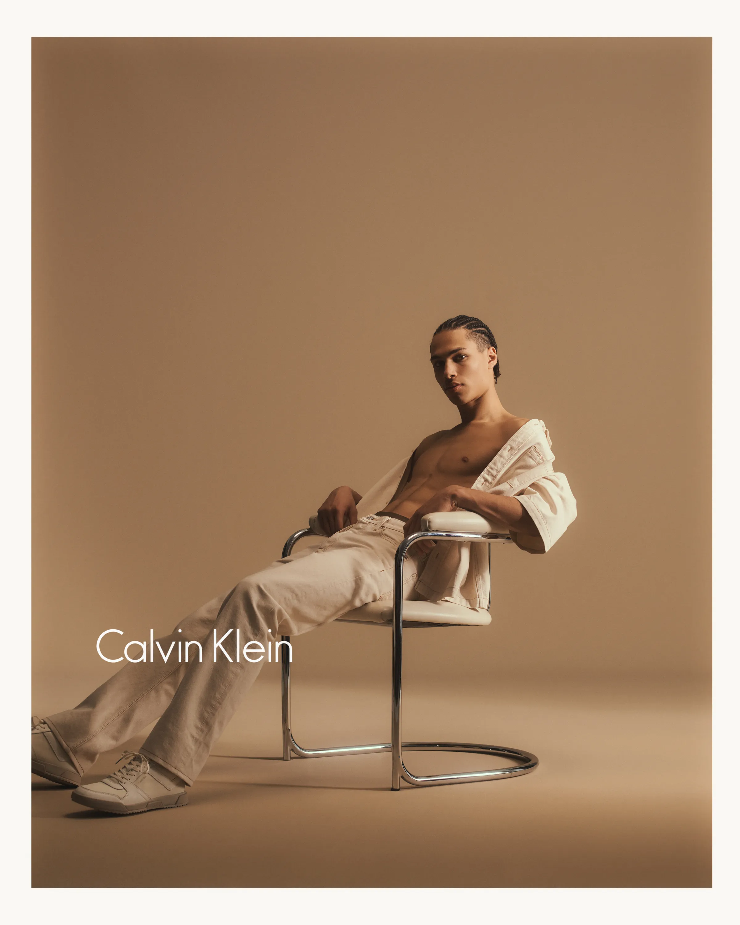Shirtless man with braided hair reclining on a modern chair wearing light pants and jacket.