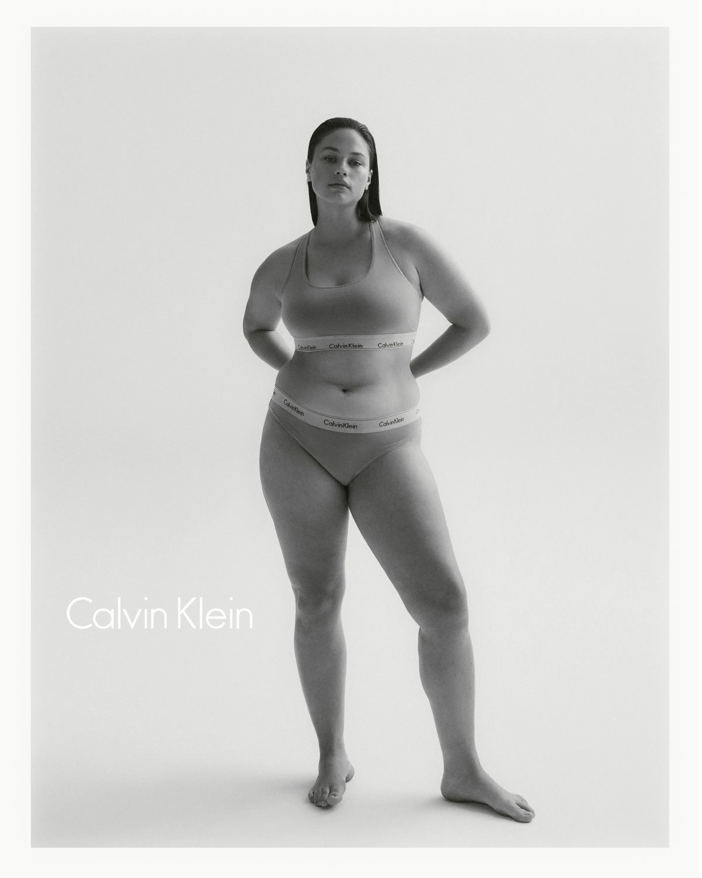 Woman standing in Calvin Klein underwear with hands behind back on plain background.