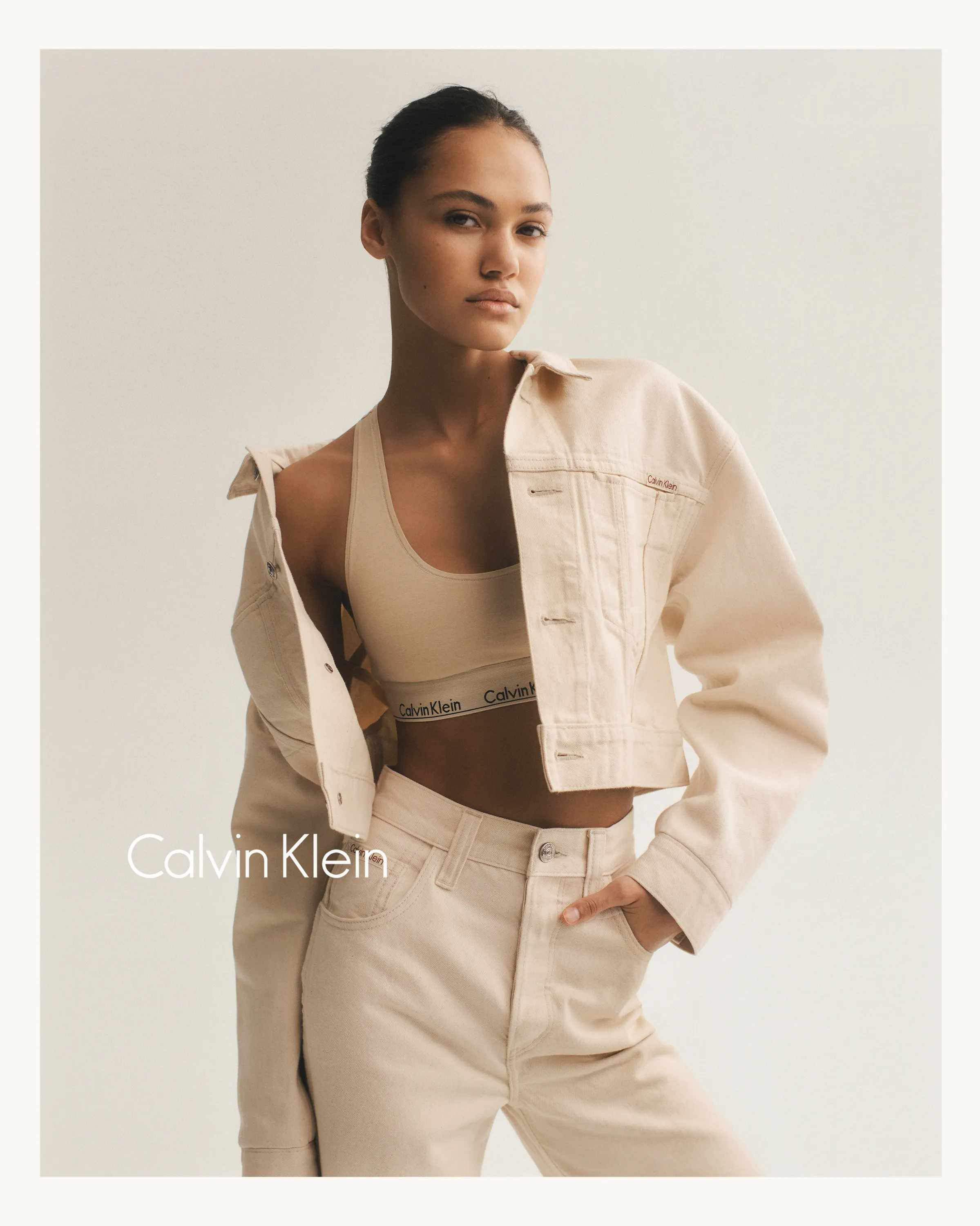 Woman in beige Calvin Klein cropped jacket, sports bra, and high-waisted pants against a plain background.