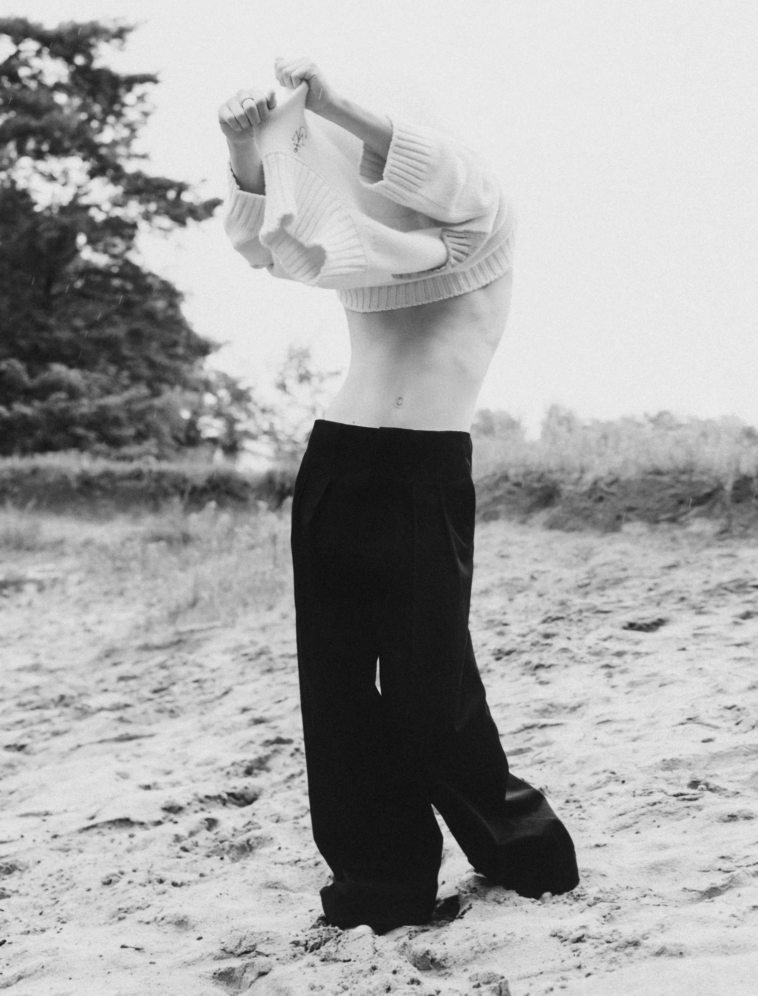 Person outdoors on sandy ground lifting a sweater over their head, showing bare midriff and wearing loose pants.