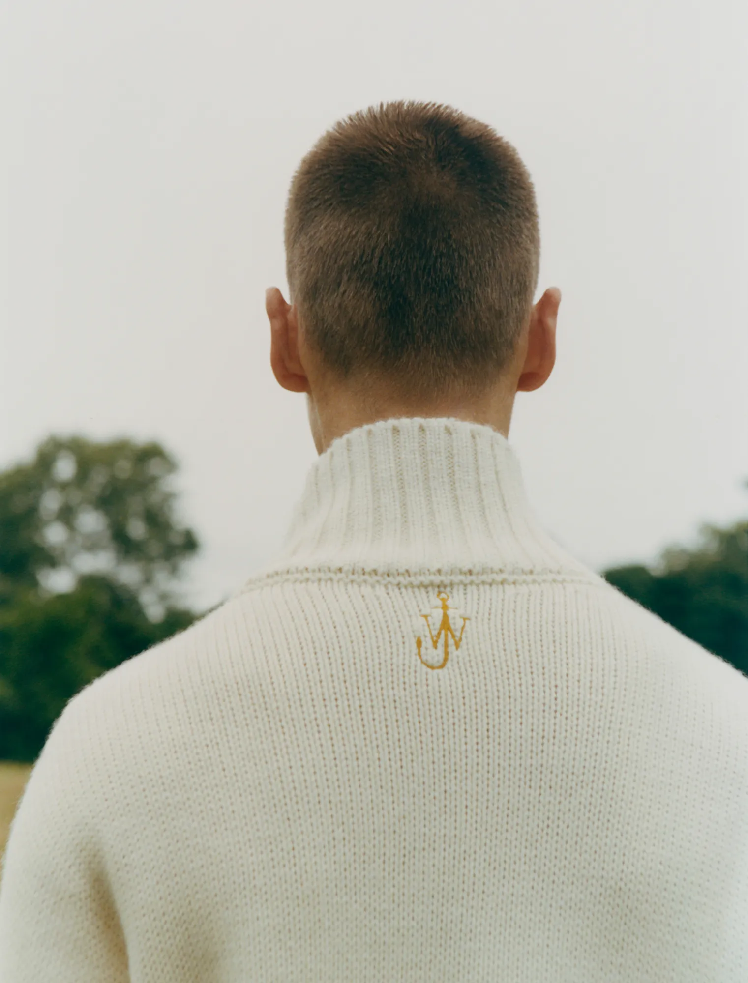 Person with short hair seen from behind wearing a cream knit sweater with a small gold emblem.