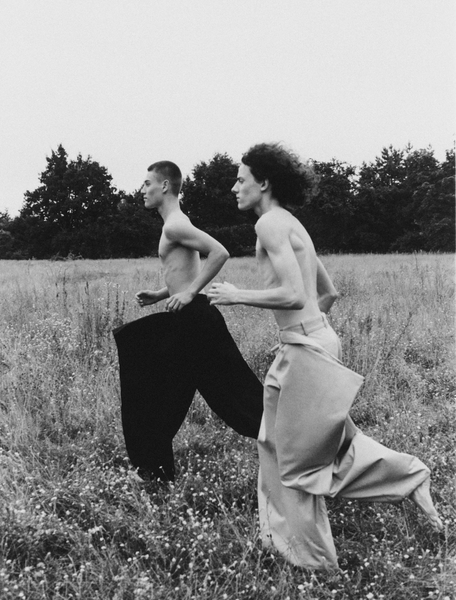 Two shirtless young men running barefoot through tall grass and wildflowers in a field.