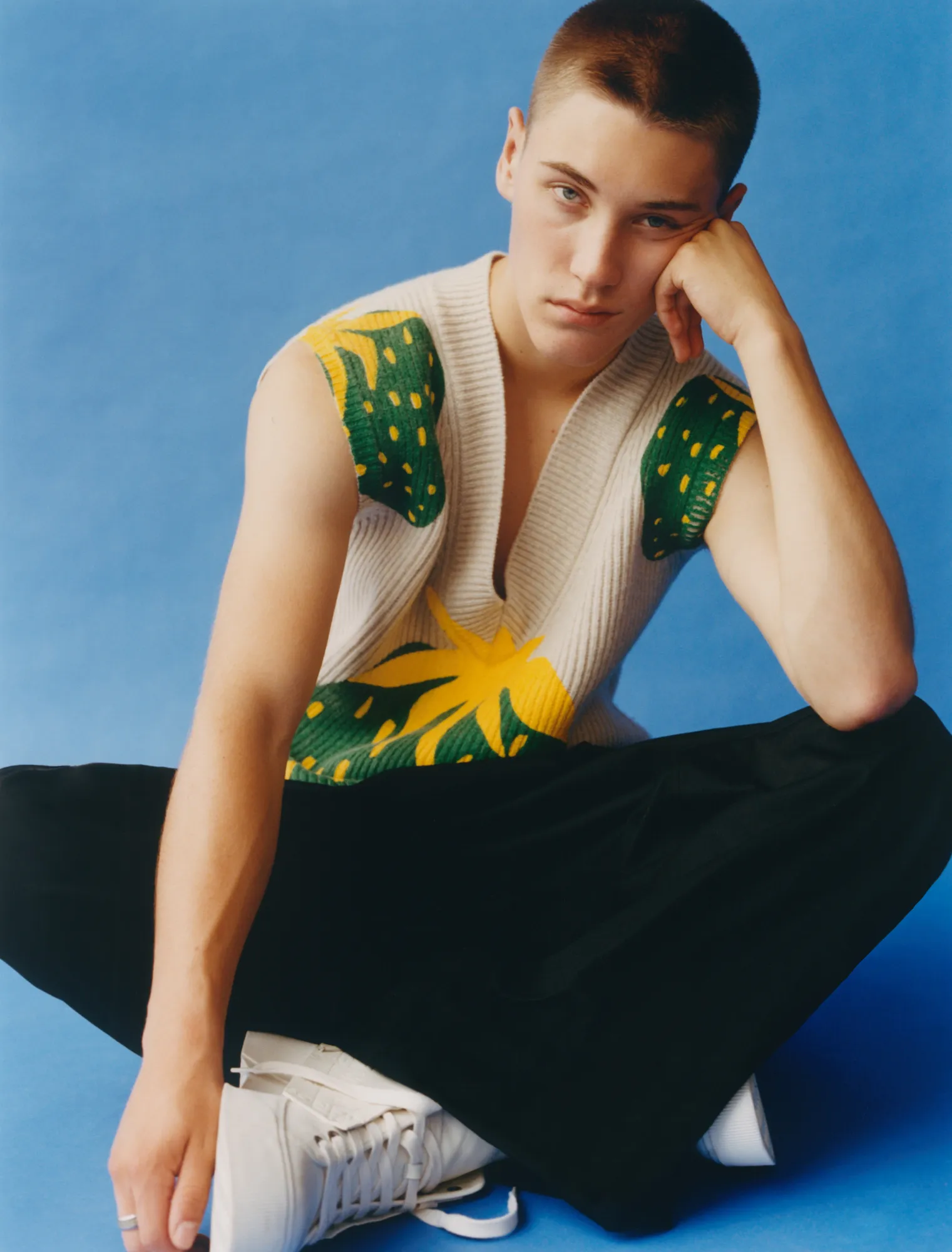 Young person with short hair wearing sleeveless sweater with green and yellow details, sitting cross-legged.