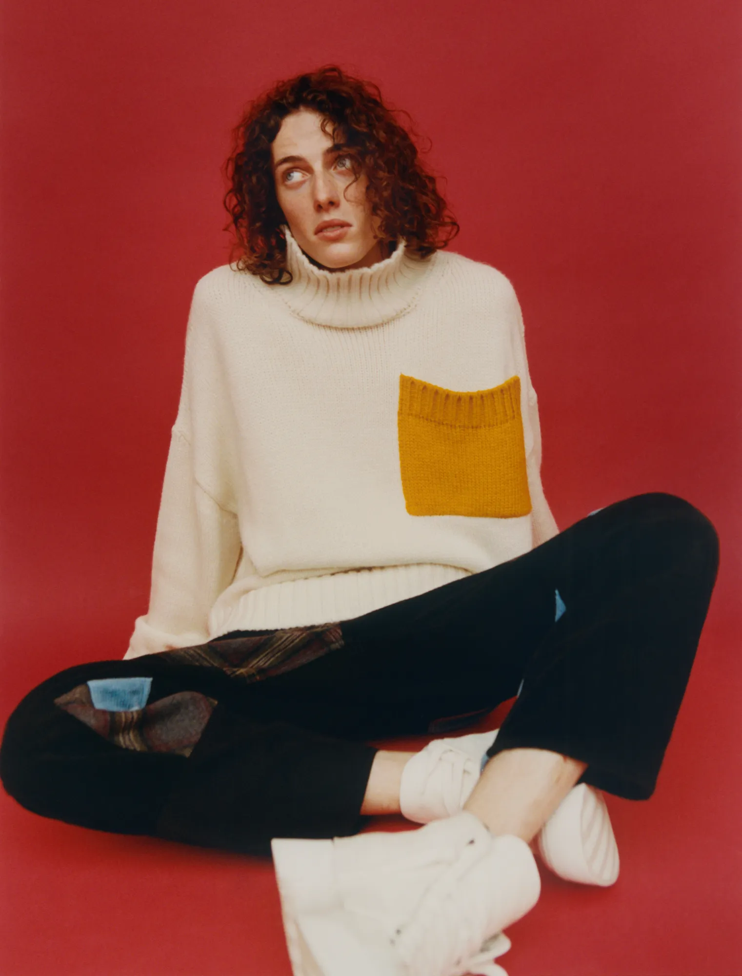 Person with curly hair wearing a cream sweater with a yellow pocket sitting on the floor against a red background.