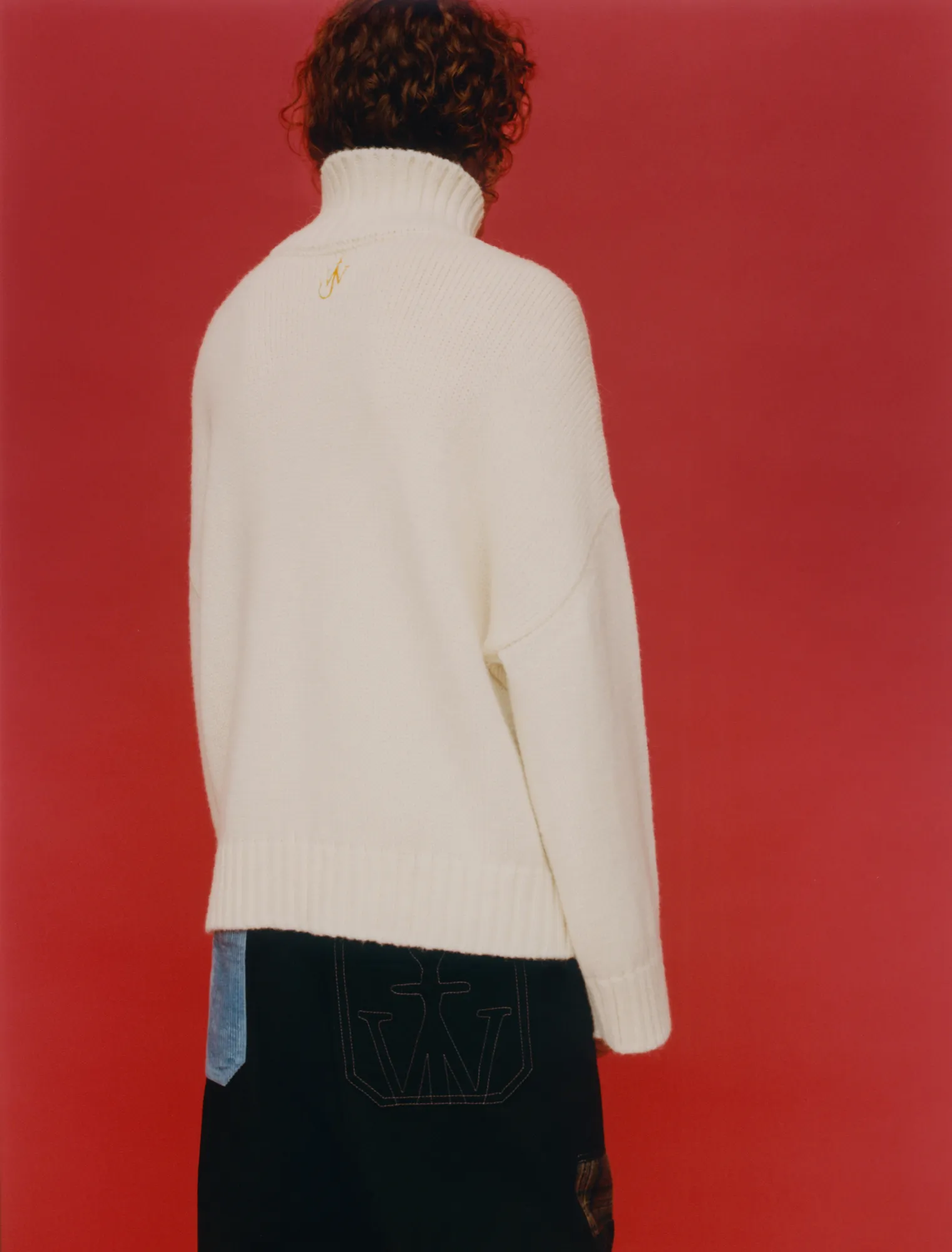 Person with curly hair wearing a white turtleneck sweater against a red background, back view.