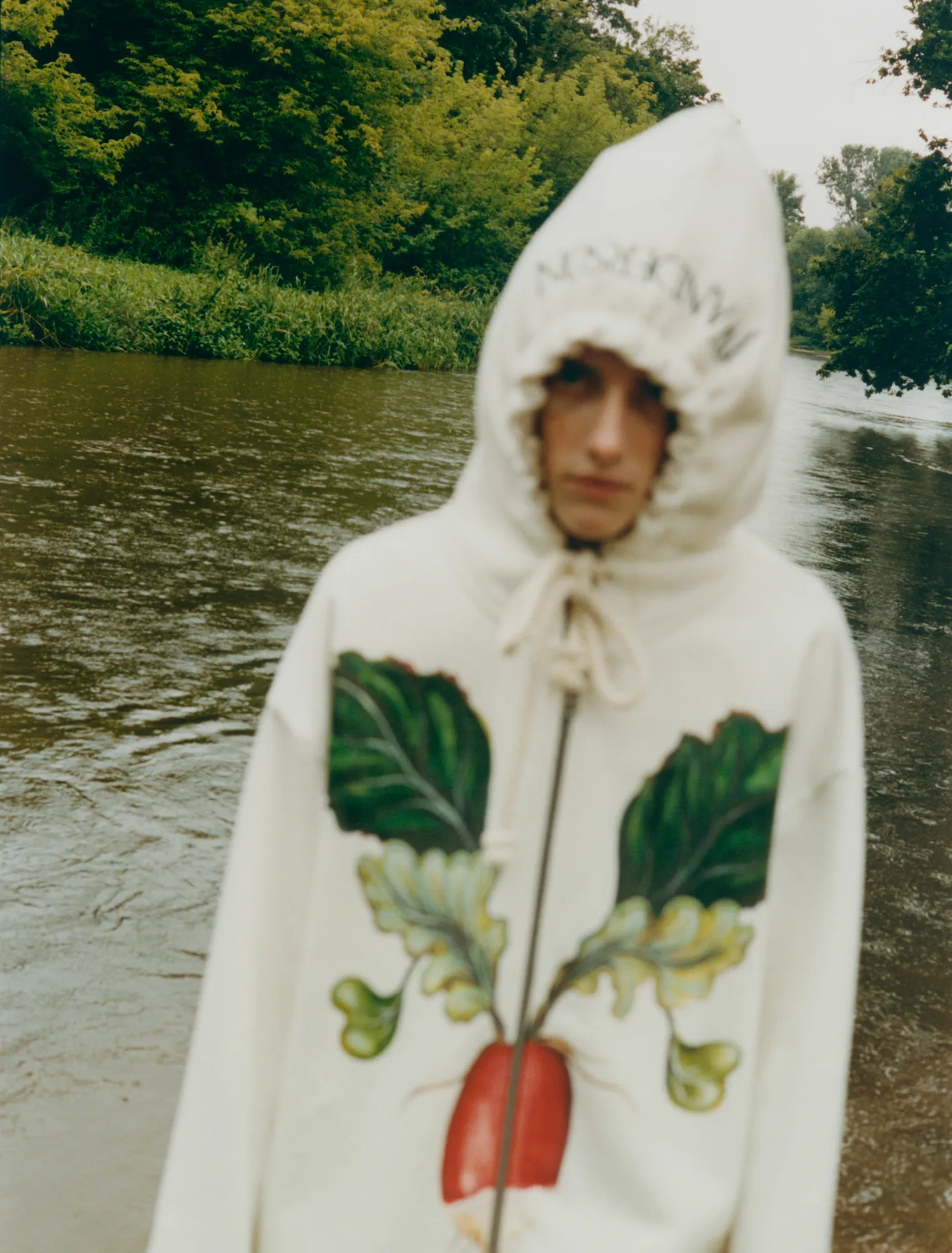 Person with hooded sweatshirt featuring a large radish print standing by a river with trees.