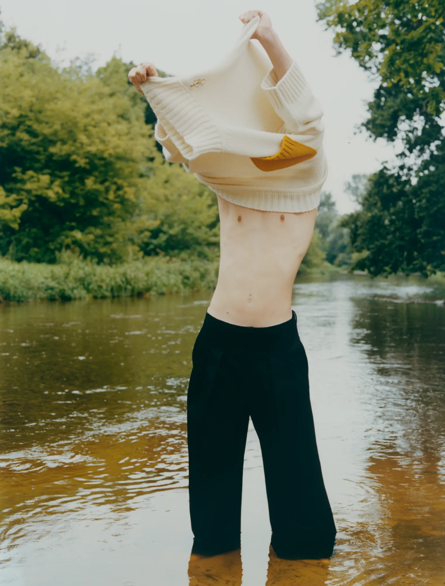 Person standing in shallow water removing a cream sweater, wearing black pants, surrounded by trees.