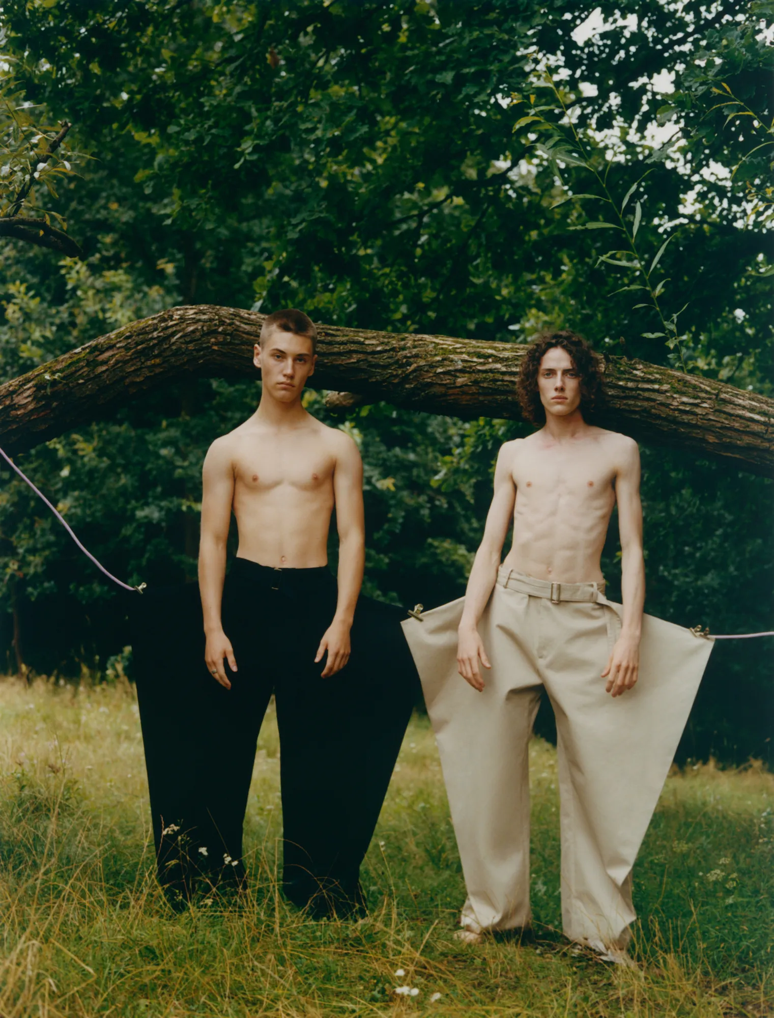 Two shirtless young men stand in oversized pants in a grassy area under a tree branch.