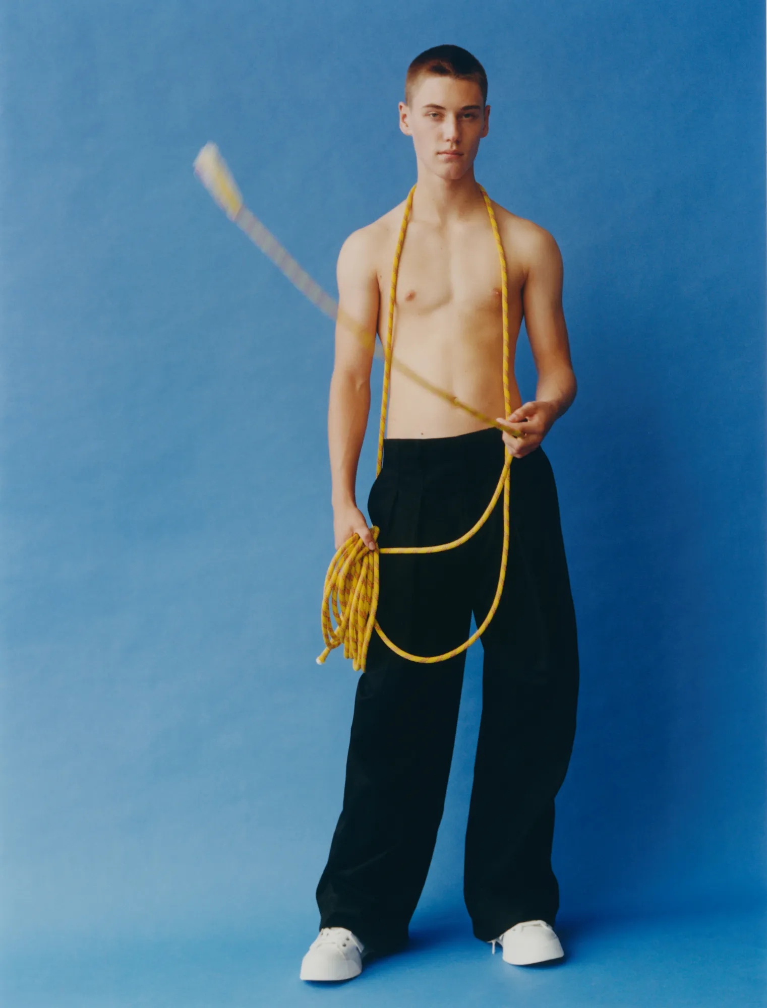 Shirtless young man holding and spinning a yellow rope against a blue background.