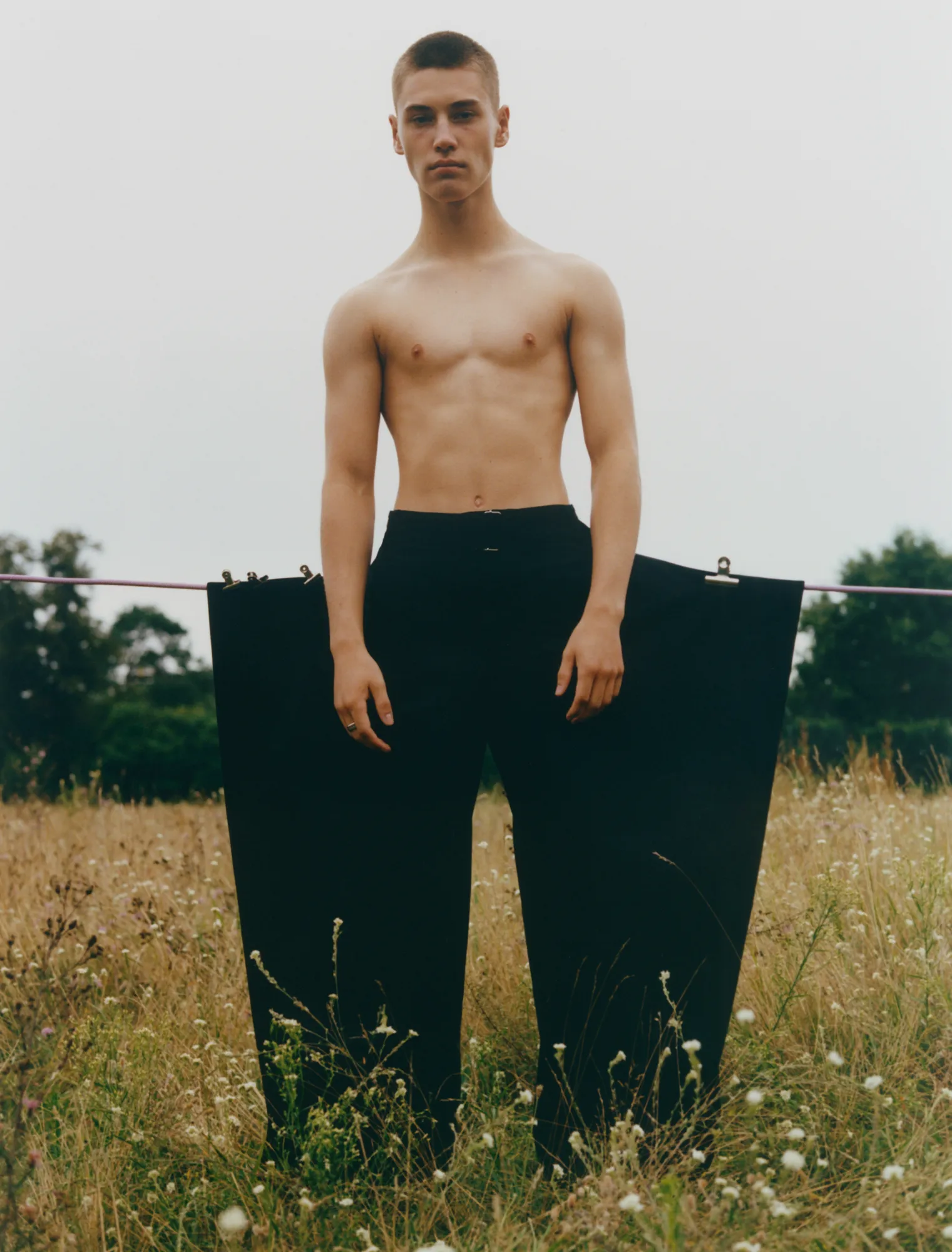 Shirtless young man stands in a field wearing oversized black pants clipped to a wire behind him.