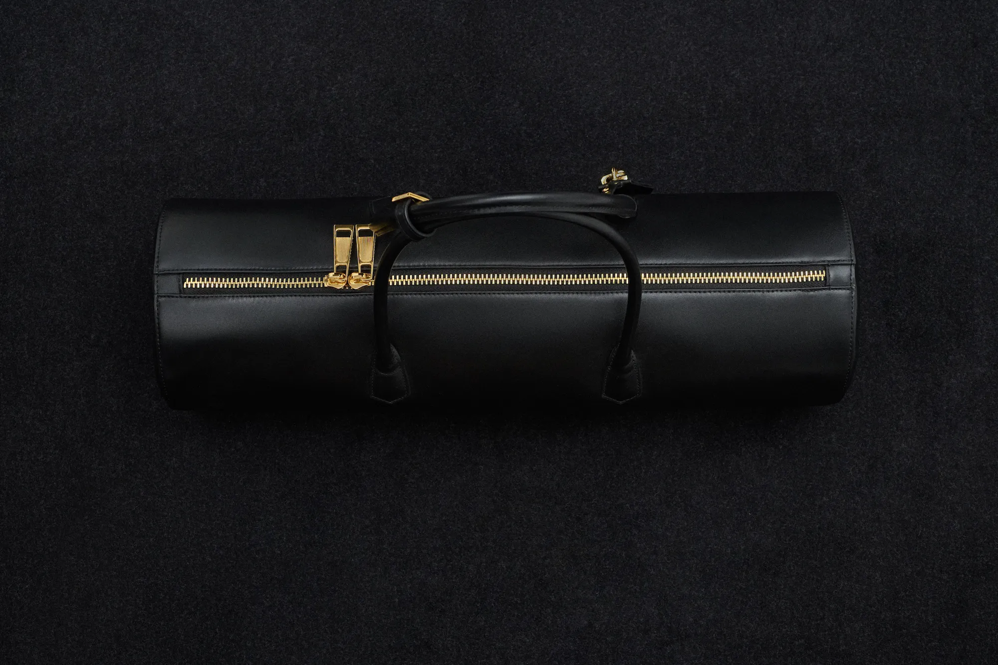Black cylindrical leather bag with gold zipper and two handles on a black textured background.