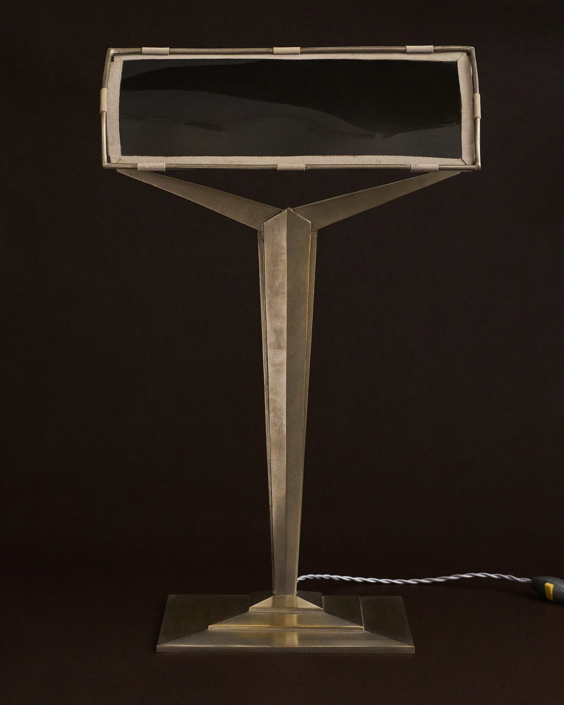Modern metal desk lamp with rectangular shade and twisted power cord on a dark background.