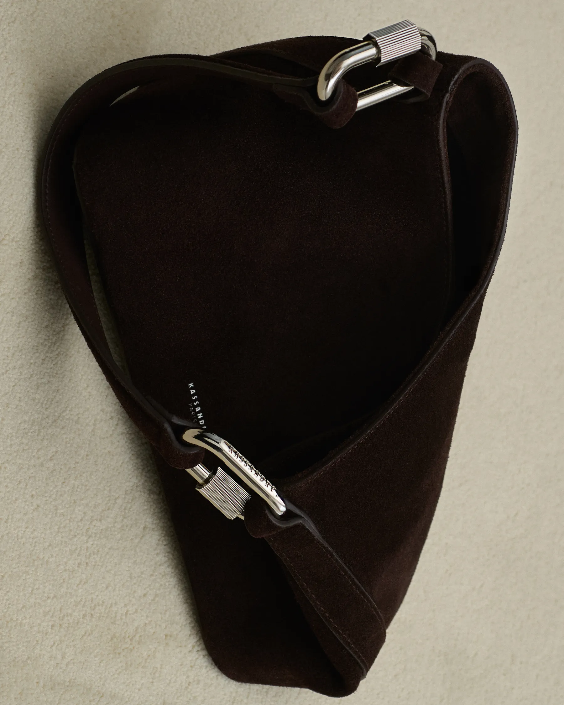 Dark brown suede shoulder bag with silver carabiner-style metal hardware on a beige background.