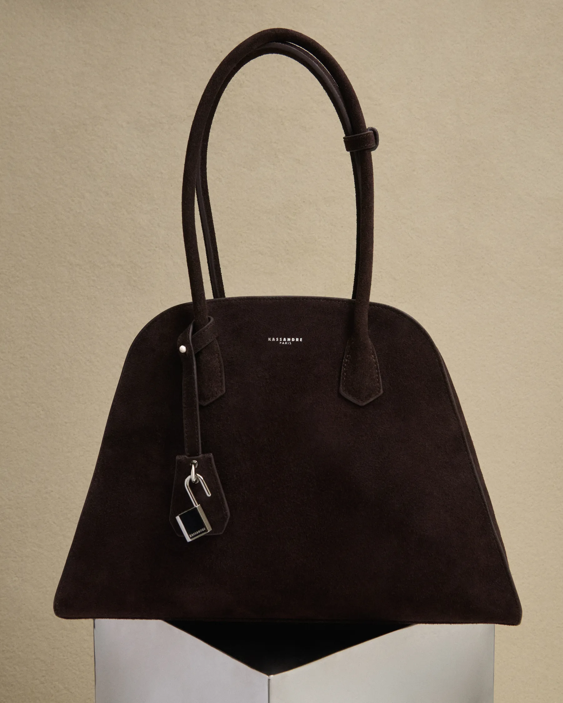 Dark brown suede handbag with two handles and a small lock accessory, displayed on a white pedestal.