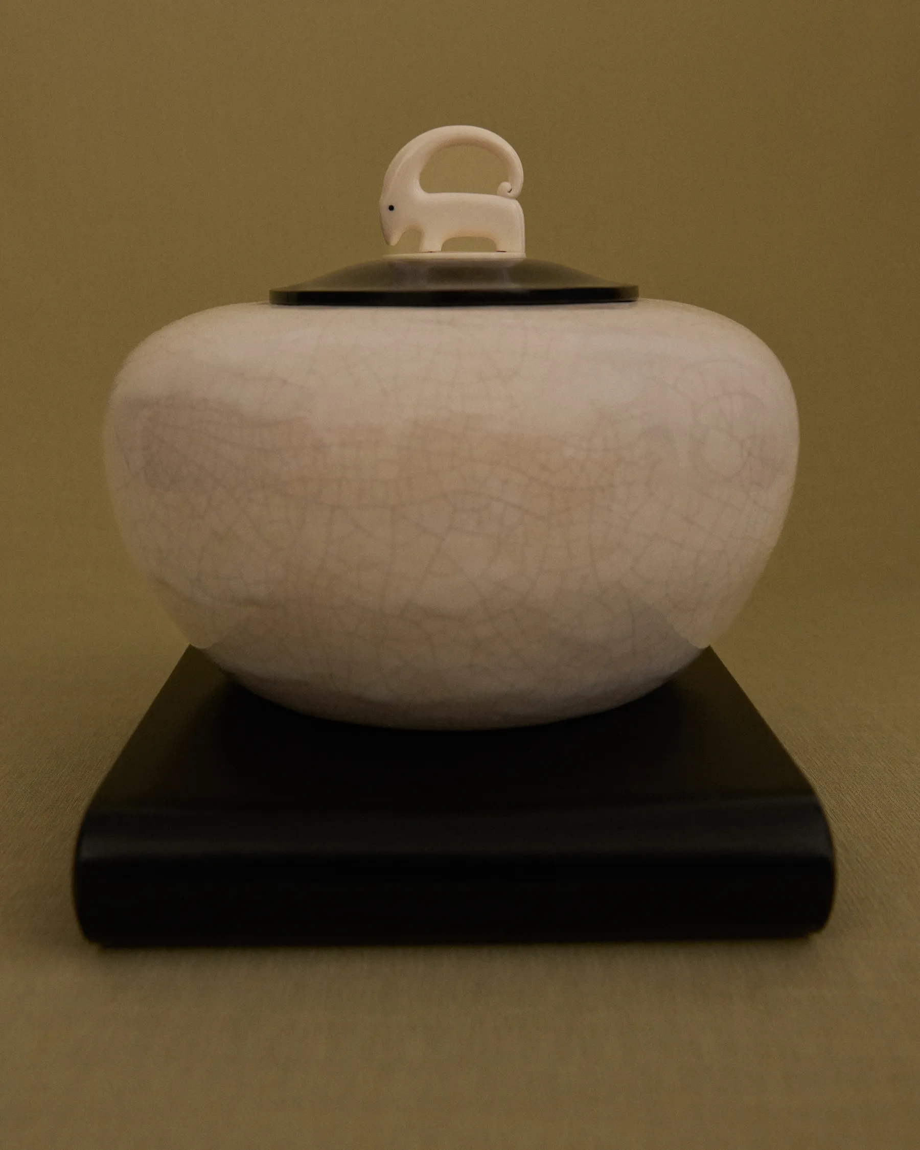 Round ceramic jar with a crackled finish and a lid topped by a small animal figurine, on a black base.