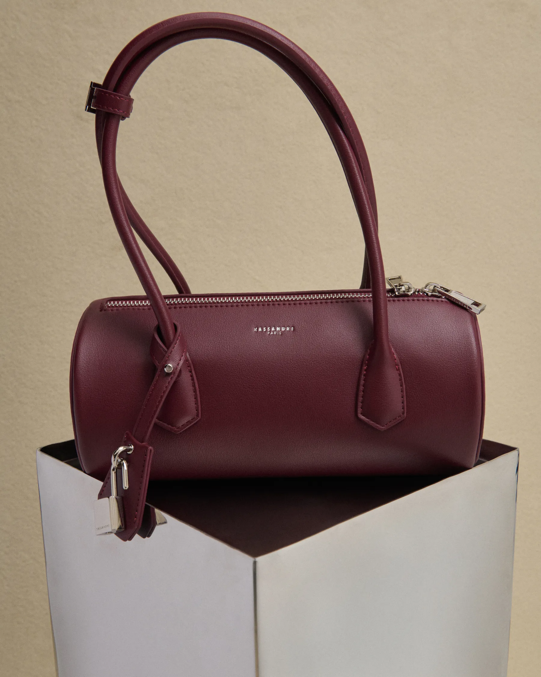 Maroon cylindrical leather handbag with double handles and silver zipper on a white pedestal.