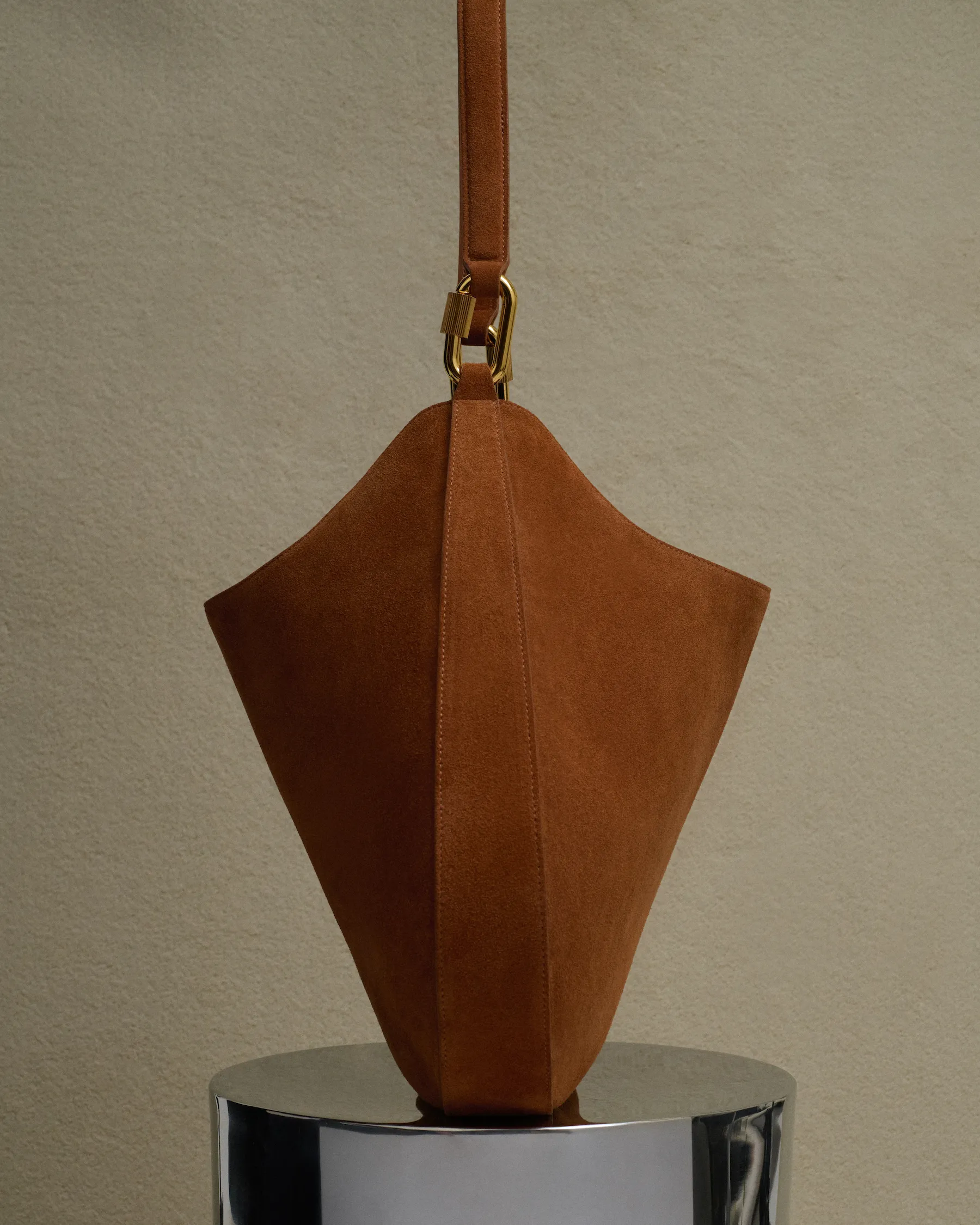 Brown suede handbag with gold hardware strap hanging above a reflective metal surface.